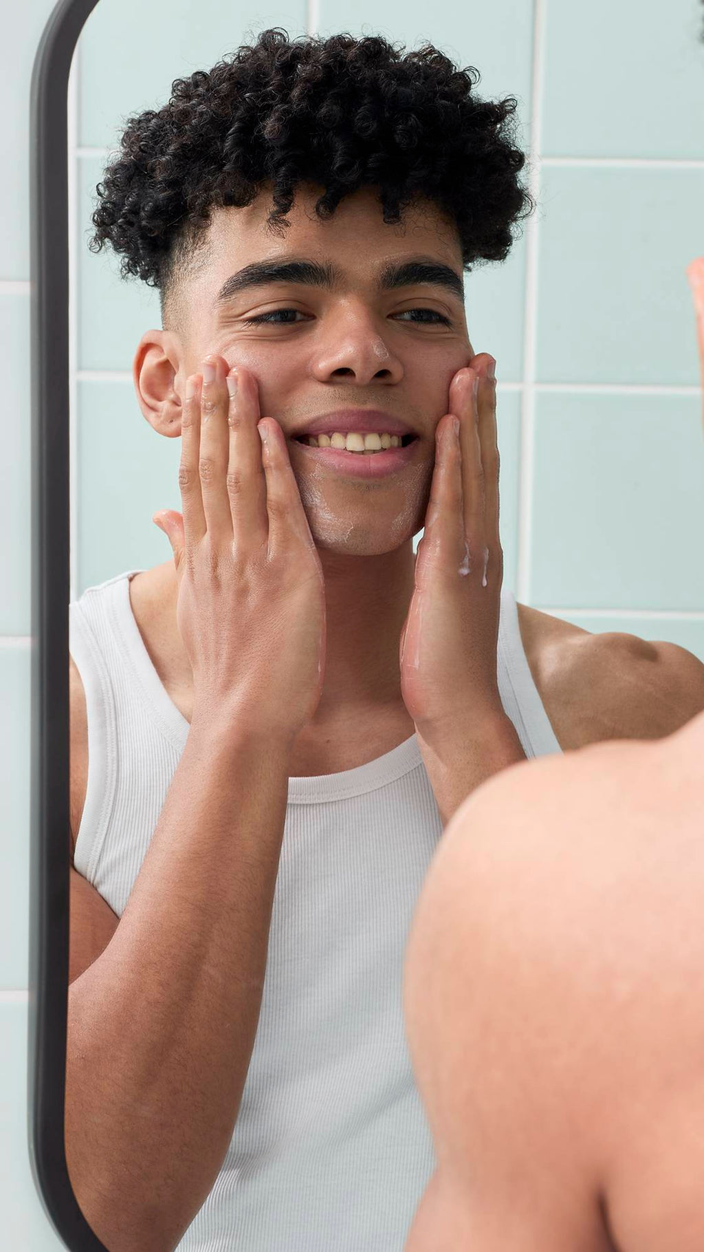 A reflection in a bathroom mirror shows a smiling model with short, curly dark hair applying a lather to their face, created by the Strawberry Picking facial gummy. They are wearing a white vest against a background of pale green wall tiles.