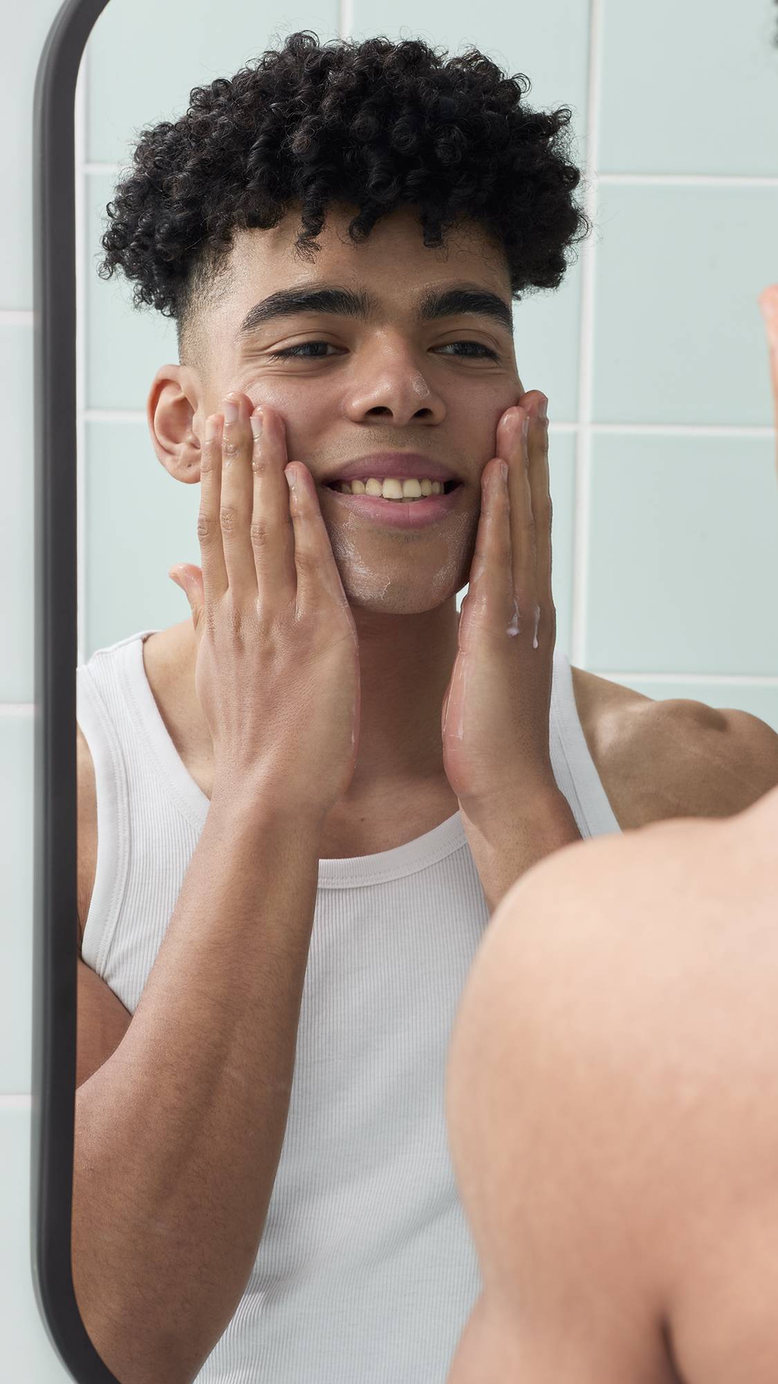 A reflection in a bathroom mirror shows a smiling model with short, curly dark hair applying a lather to their face, created by the Strawberry Picking facial gummy. They are wearing a white vest against a background of pale green wall tiles.