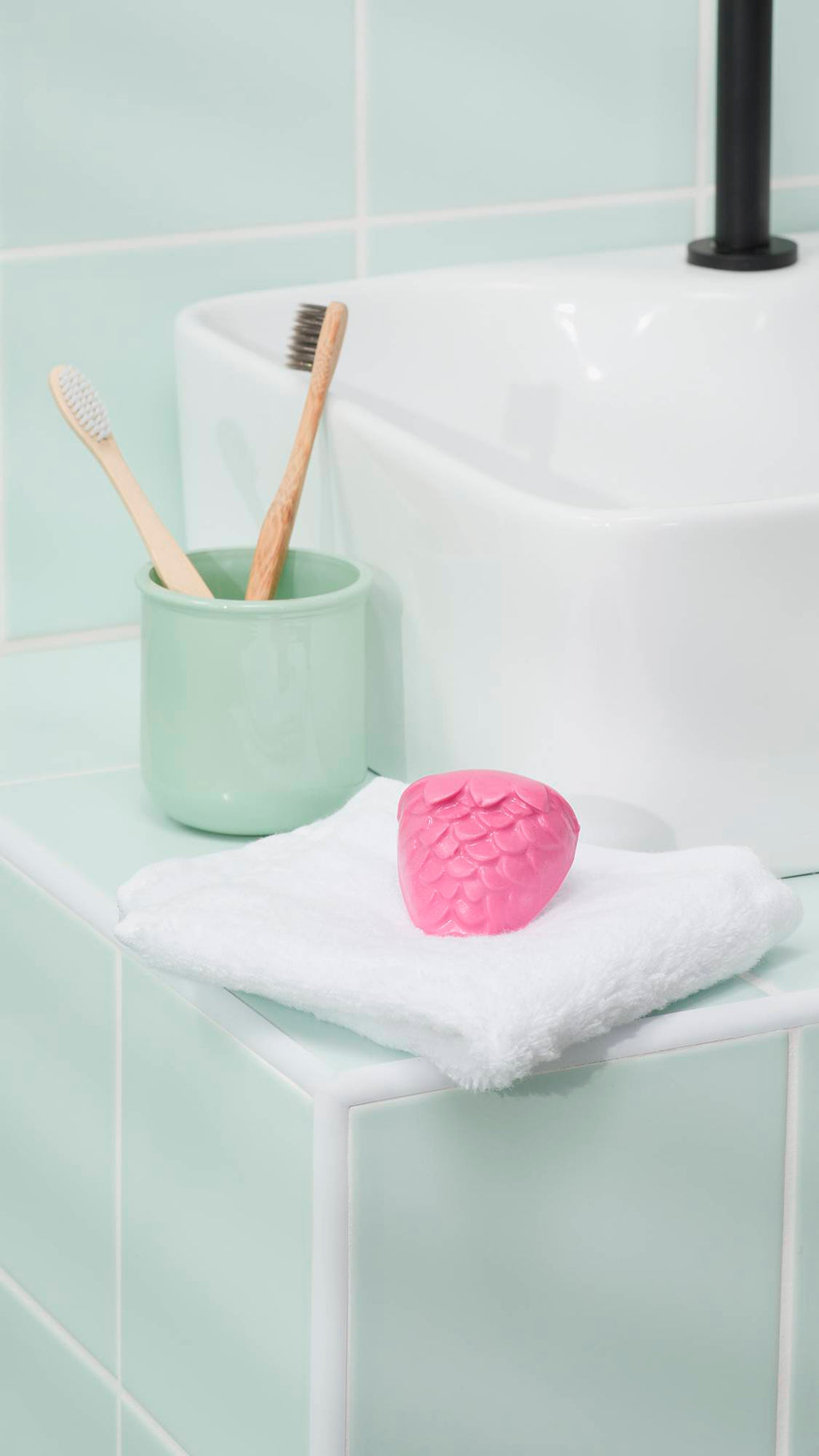 A vibrant pink "Strawberry Picking" facial gummy sits on a folded white towel on a mint green tiled bathroom counter. In the background, two bamboo toothbrushes rest in a matching green ceramic pot next to a white ceramic sink with a sleek black tap.