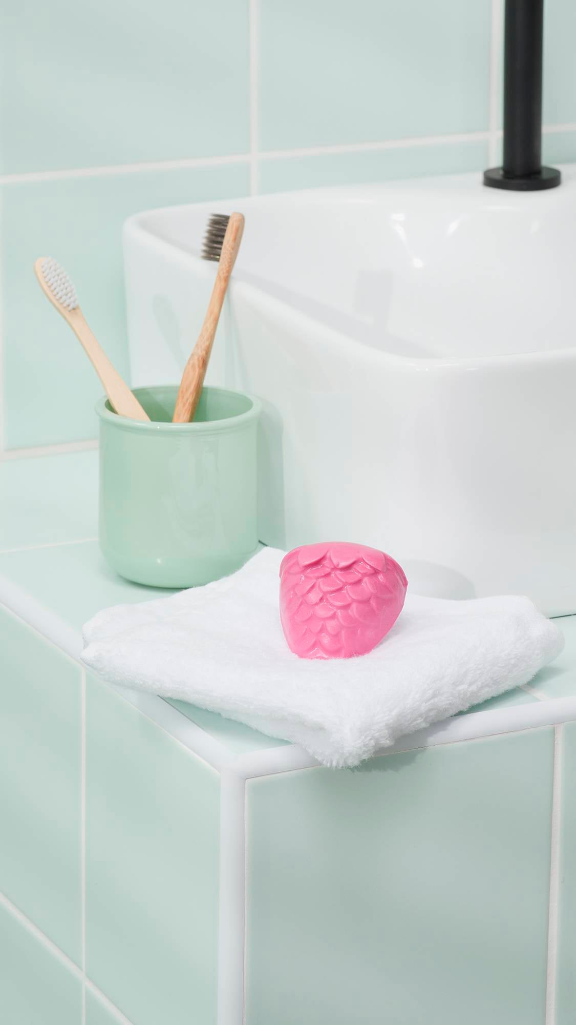 A vibrant pink "Strawberry Picking" facial gummy sits on a folded white towel on a mint green tiled bathroom counter. In the background, two bamboo toothbrushes rest in a matching green ceramic pot next to a white ceramic sink with a sleek black tap.