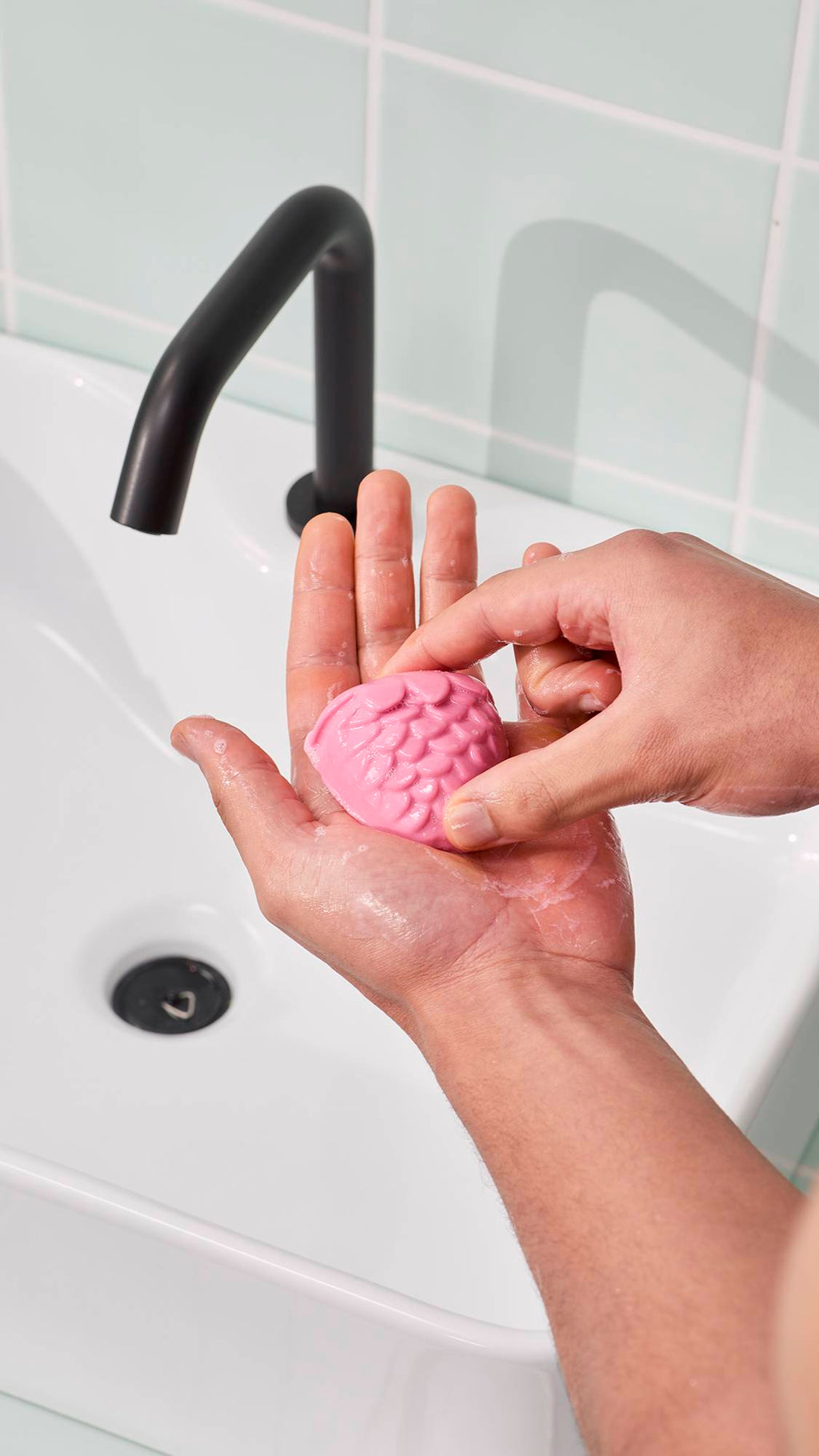 A model holds a pink, strawberry-shaped facial jelly in their hands to create a lather. The "Strawberry Picking" facial jelly cleanser has a scalloped texture. In the background, a white sink and black tap are visible against pale green wall tiles.