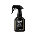 A recycled black plastic LUSH body spray bottle with a trigger nozzle stands upright with a creamy-yellow rimmed label with white text that reads "Super Milk Body Spray".
