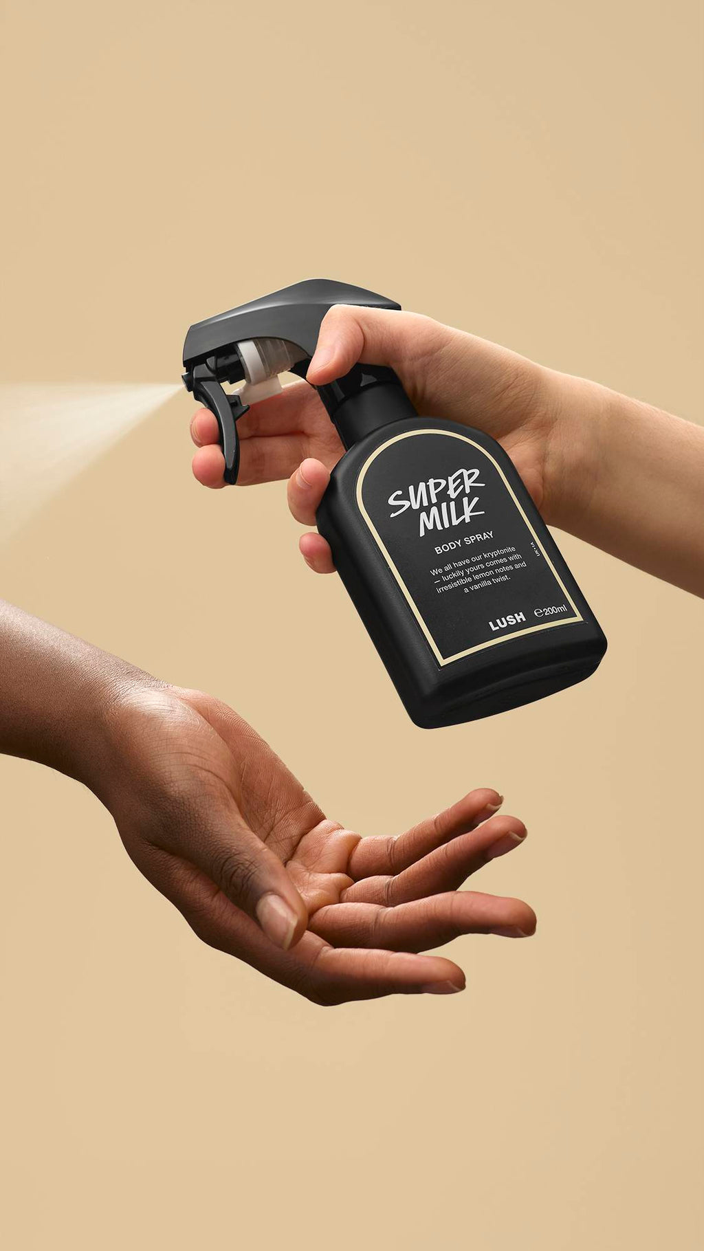 The image shows a light-toned hand spraying a bottle of LUSH 'Super Milk' body spray. A warm-toned hand is cupped below, catching the fine mist against a warm, beige background.
