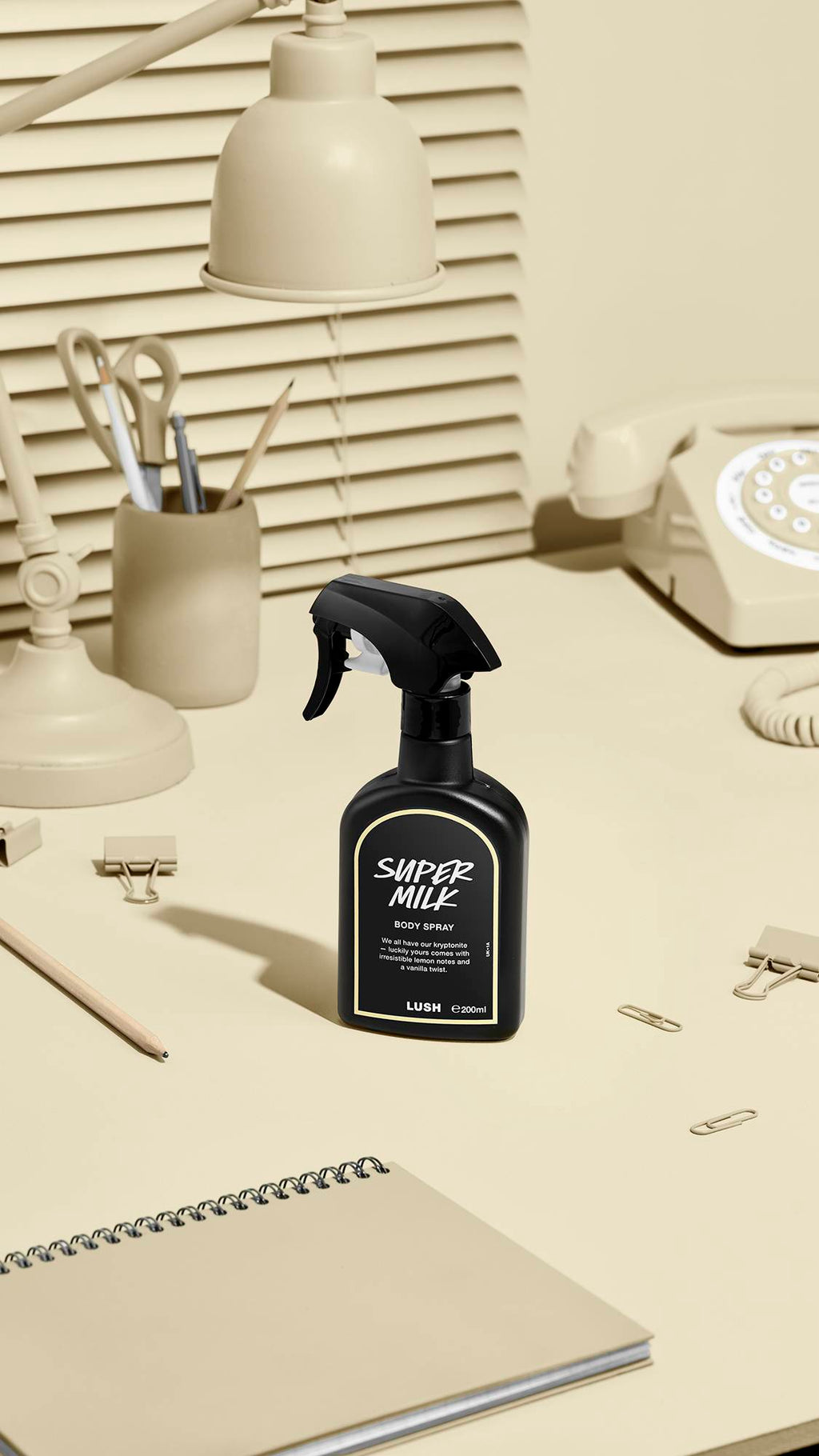 The image shows a black LUSH 'Super Milk' body spray bottle with a pale beige label on a beige desk. Nearby sit a matching desk lamp, a rotary phone, stationery, and a notebook, against window blinds.