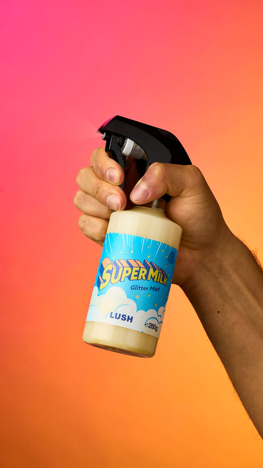 A hand holds and sprays a bottle of Lush 'Super Milk' Glitter Mist, releasing a fine vapour. The bottle has a black spray nozzle, a bright blue cloud-patterned label, and is held against a vibrant pink-to-orange gradient background.