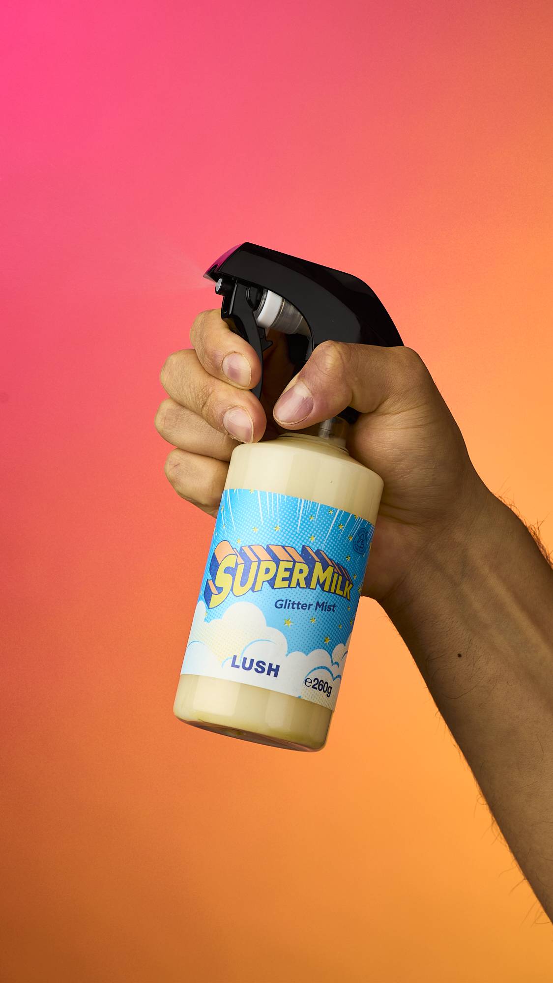 A hand holds and sprays a bottle of Lush 'Super Milk' Glitter Mist, releasing a fine vapour. The bottle has a black spray nozzle, a bright blue cloud-patterned label, and is held against a vibrant pink-to-orange gradient background.