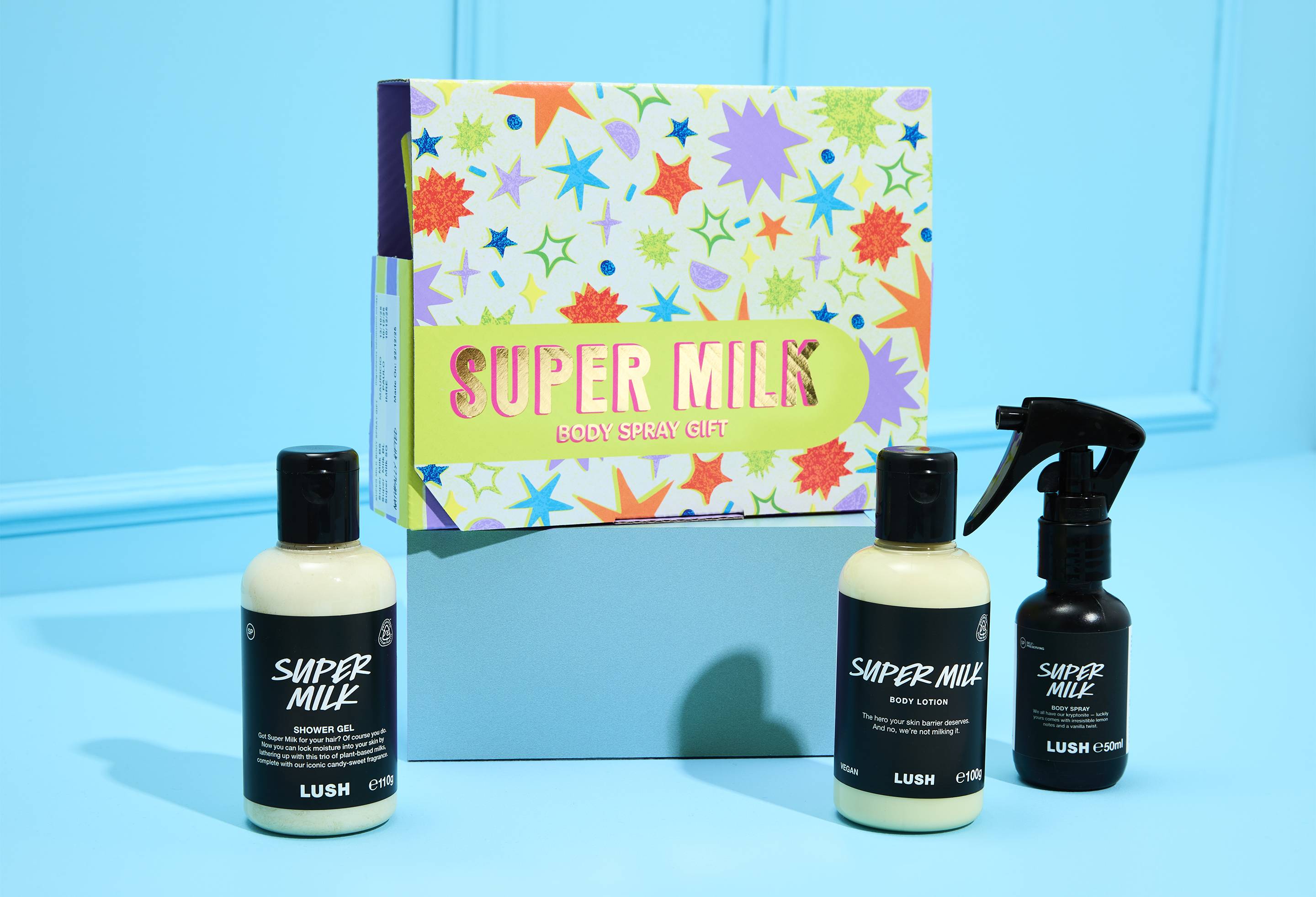 The Super Milk Body Spray Gift set sits on a blue surface. A colourful star-patterned box stands behind bottles of Shower Gel, Body Lotion, and Body Spray.