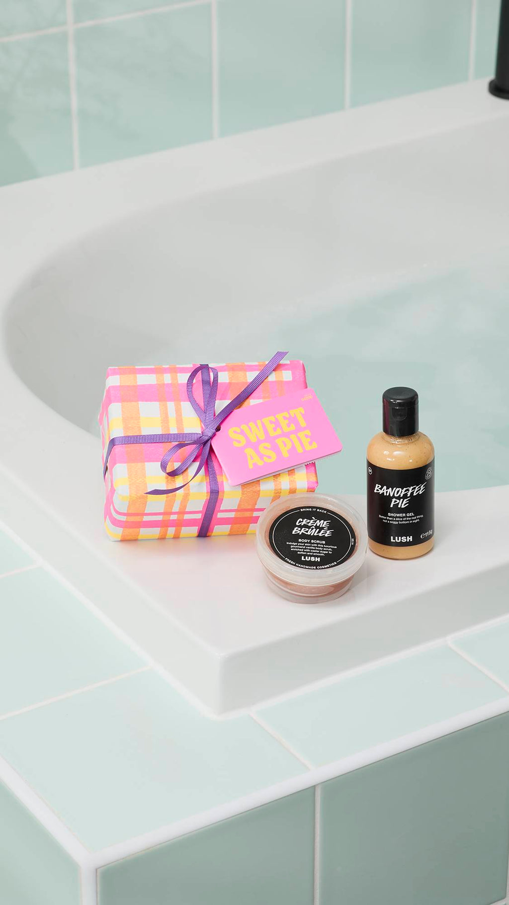 A "Sweet As Pie" gift set is displayed on the edge of a white bathtub. It includes a pink and orange checkered gift box with a purple ribbon, a pot of Crème Brûlée body scrub, and a bottle of Banoffee Pie shower gel against light green tiles.
