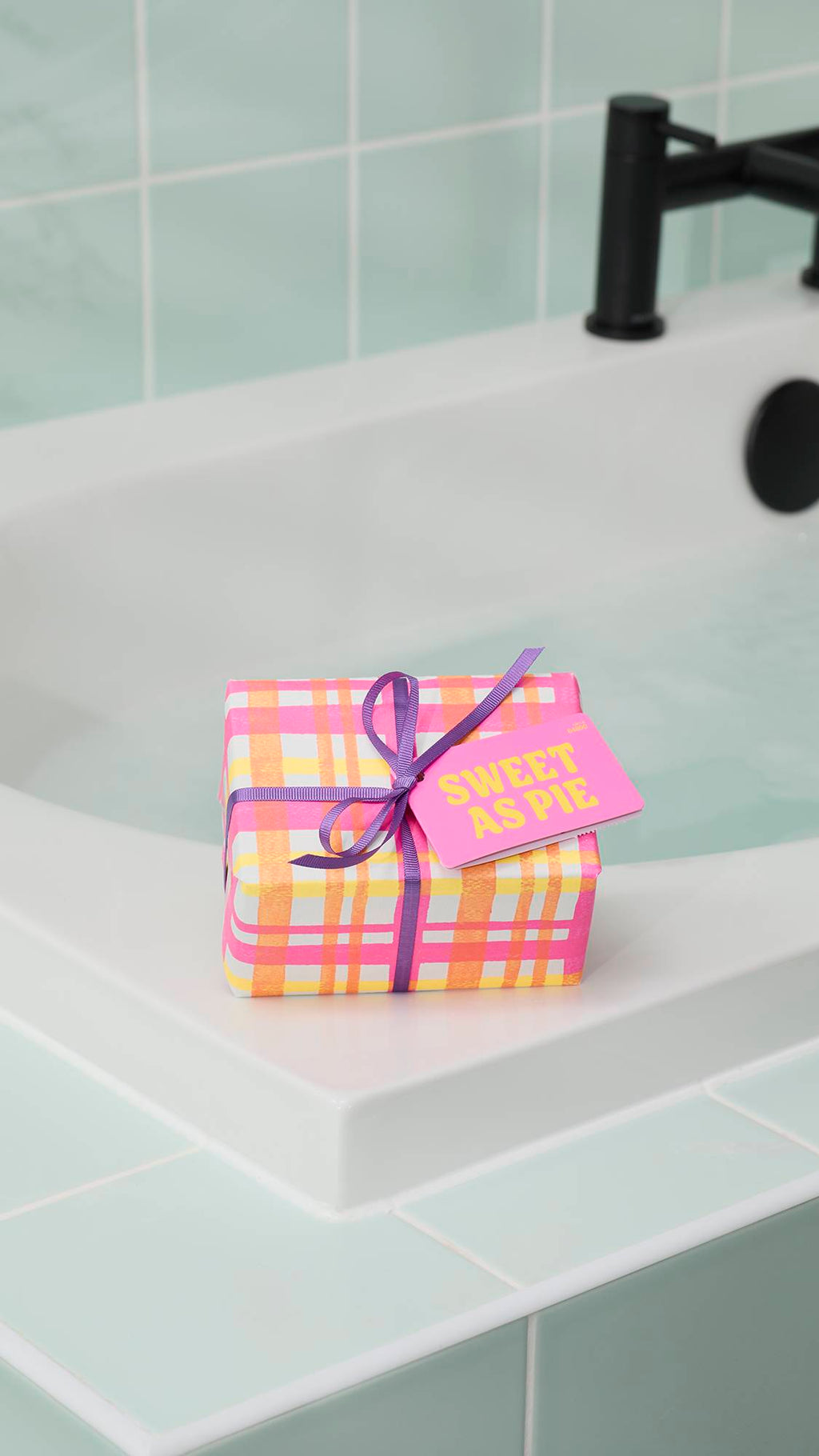 The "Sweet As Pie" gift set, wrapped in a pink and orange checkered pattern with a purple ribbon and bright pink tag, sits on the white corner of a bathtub. The background features pale green wall tiles and a black tap.