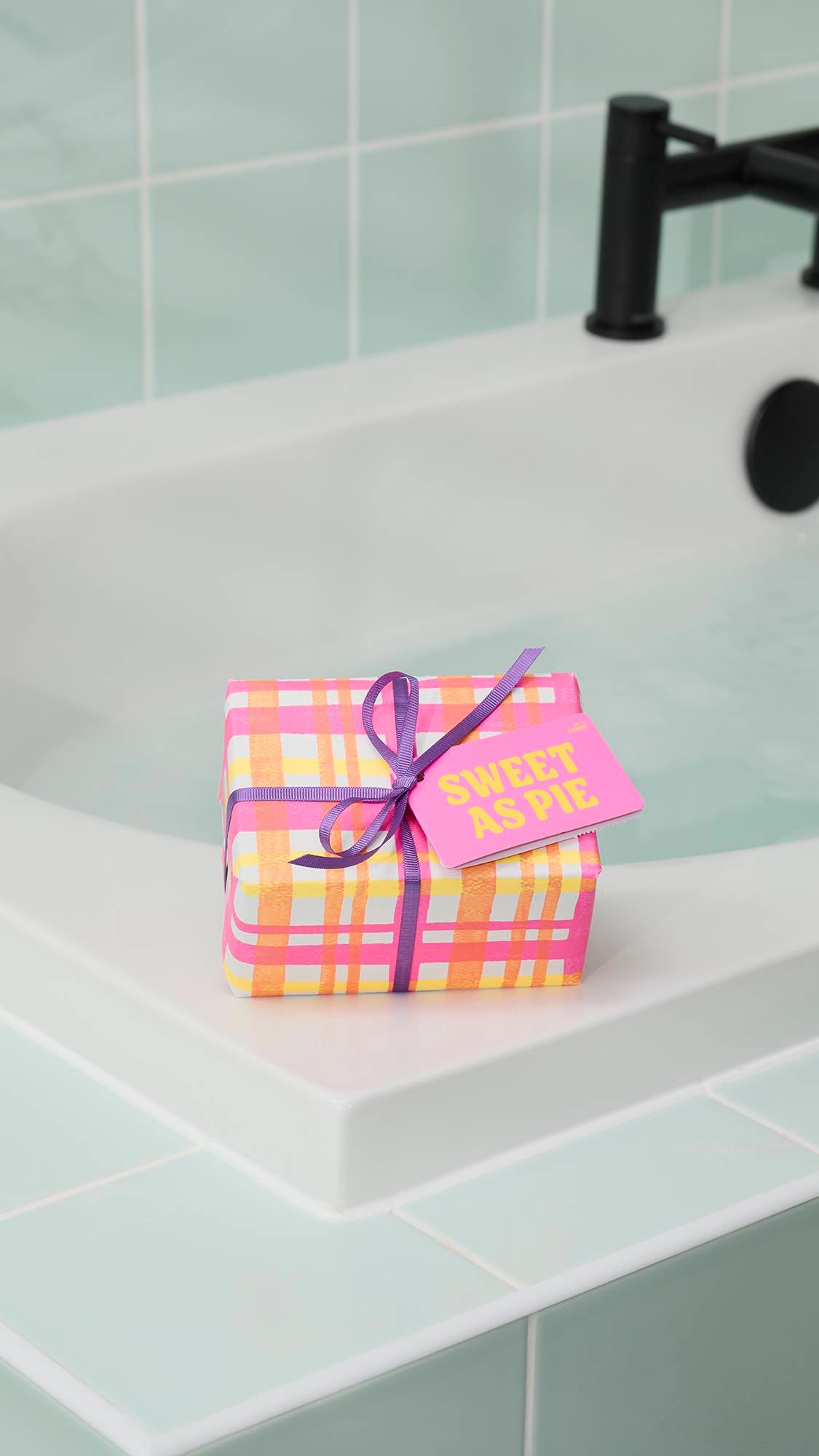 The "Sweet As Pie" gift set, wrapped in a pink and orange checkered pattern with a purple ribbon and bright pink tag, sits on the white corner of a bathtub. The background features pale green wall tiles and a black tap.