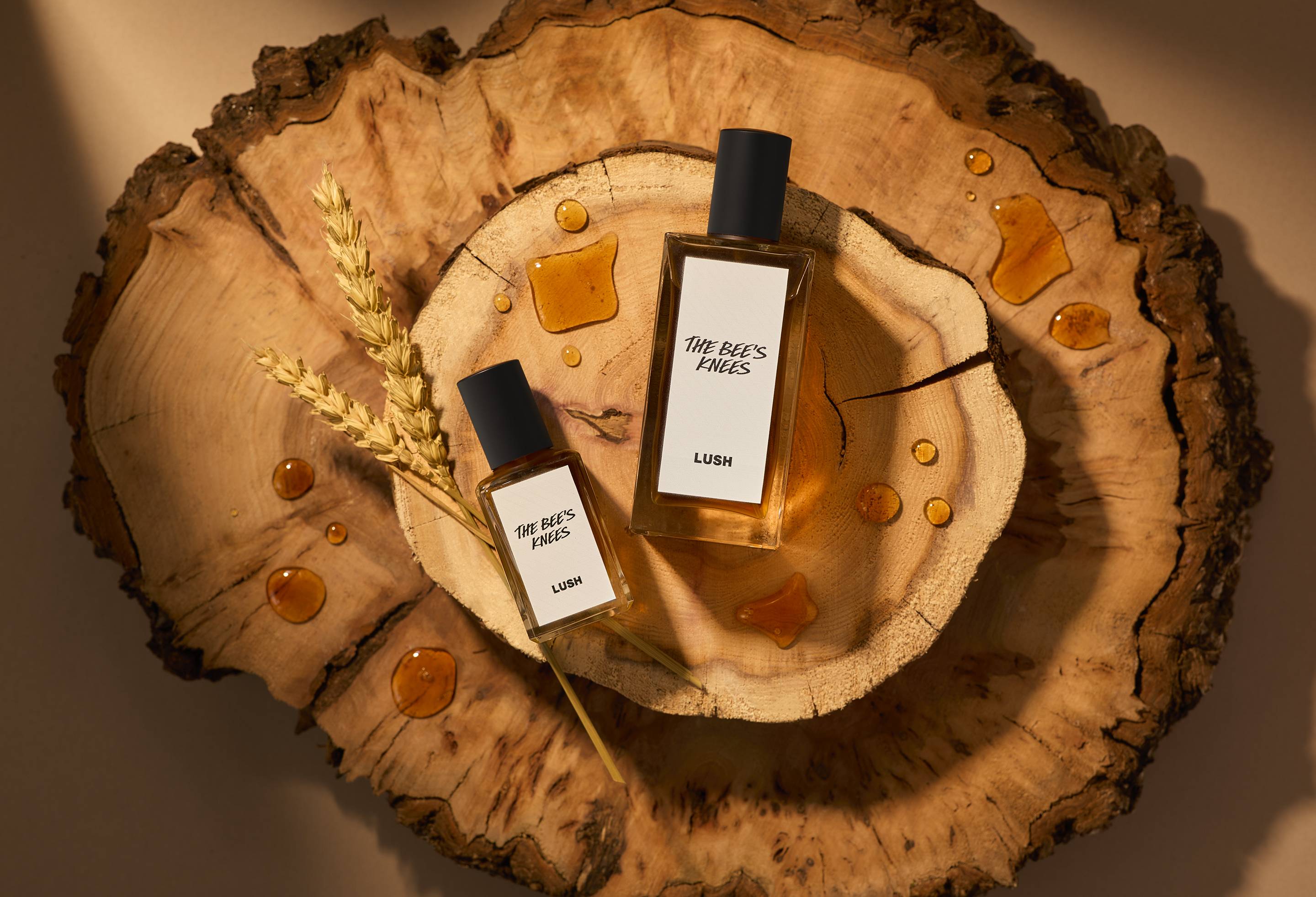 The image shows two stacked slices of wood. There is a warm light beaming over the two sizes of perfume bottles laid flat next to strands of wheat and dripped with splashes of honey.