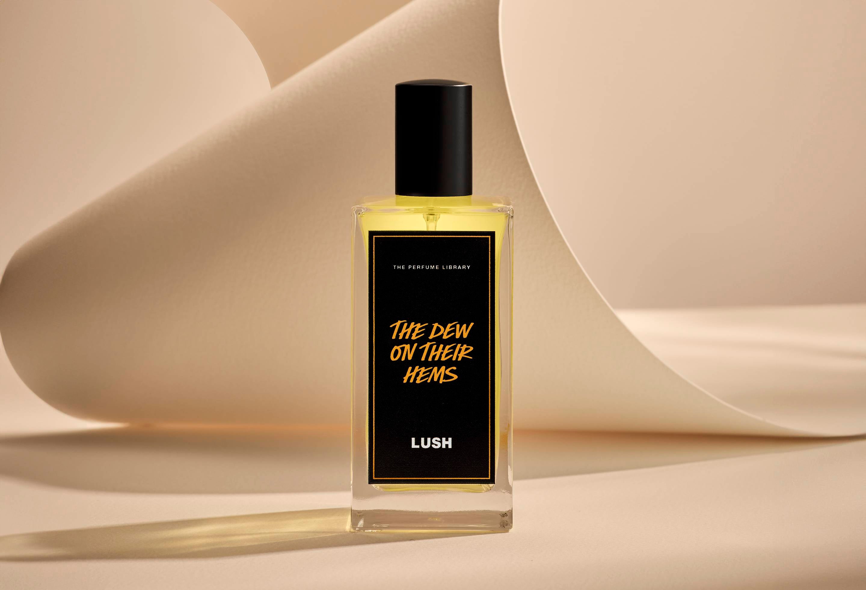 The image shows The Dew On Their Hems glass perfume bottle. It is sitting on a soft, beige background with curved, cream paper creating soft, gliding shapes behind the product.
