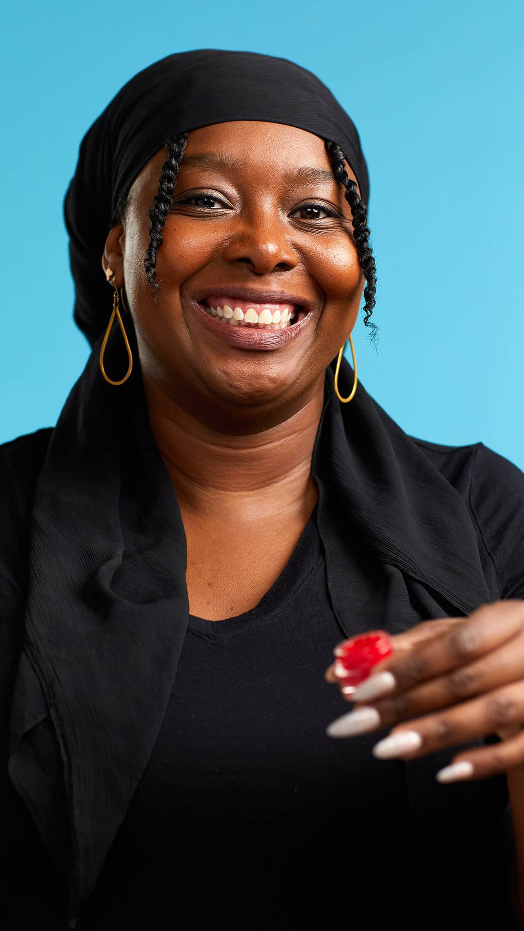 A close-up shot of the model on a sky-blue background wearing a black top. They are holding a small, glass pot of The Kiss lip gloss with sparkling, elegant nails as they smile at the camera.