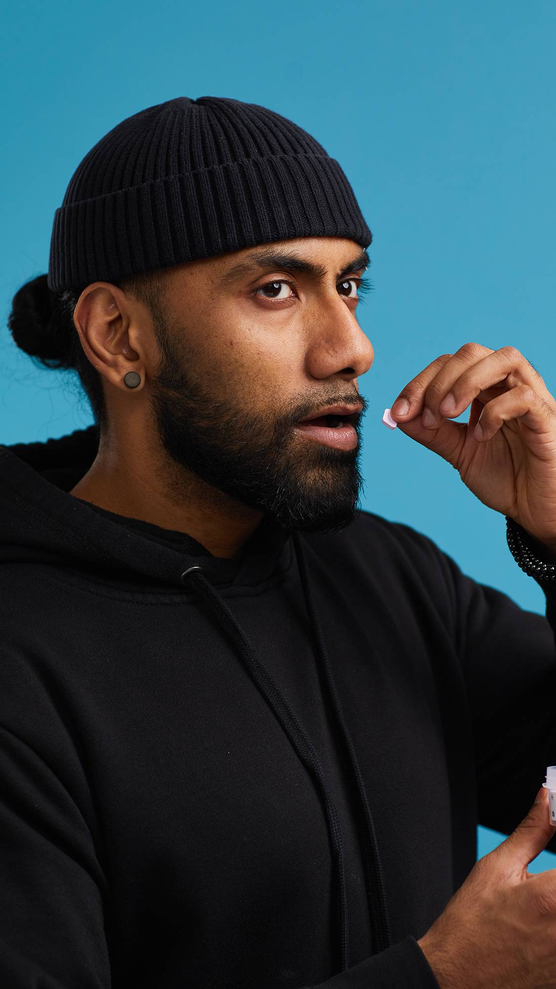 The image shows the model on a sky blue background wearing a back hoodie and hat. They are holding a small, pink The Kiss mouthwash tablet in front of their face, their mouth just opening.