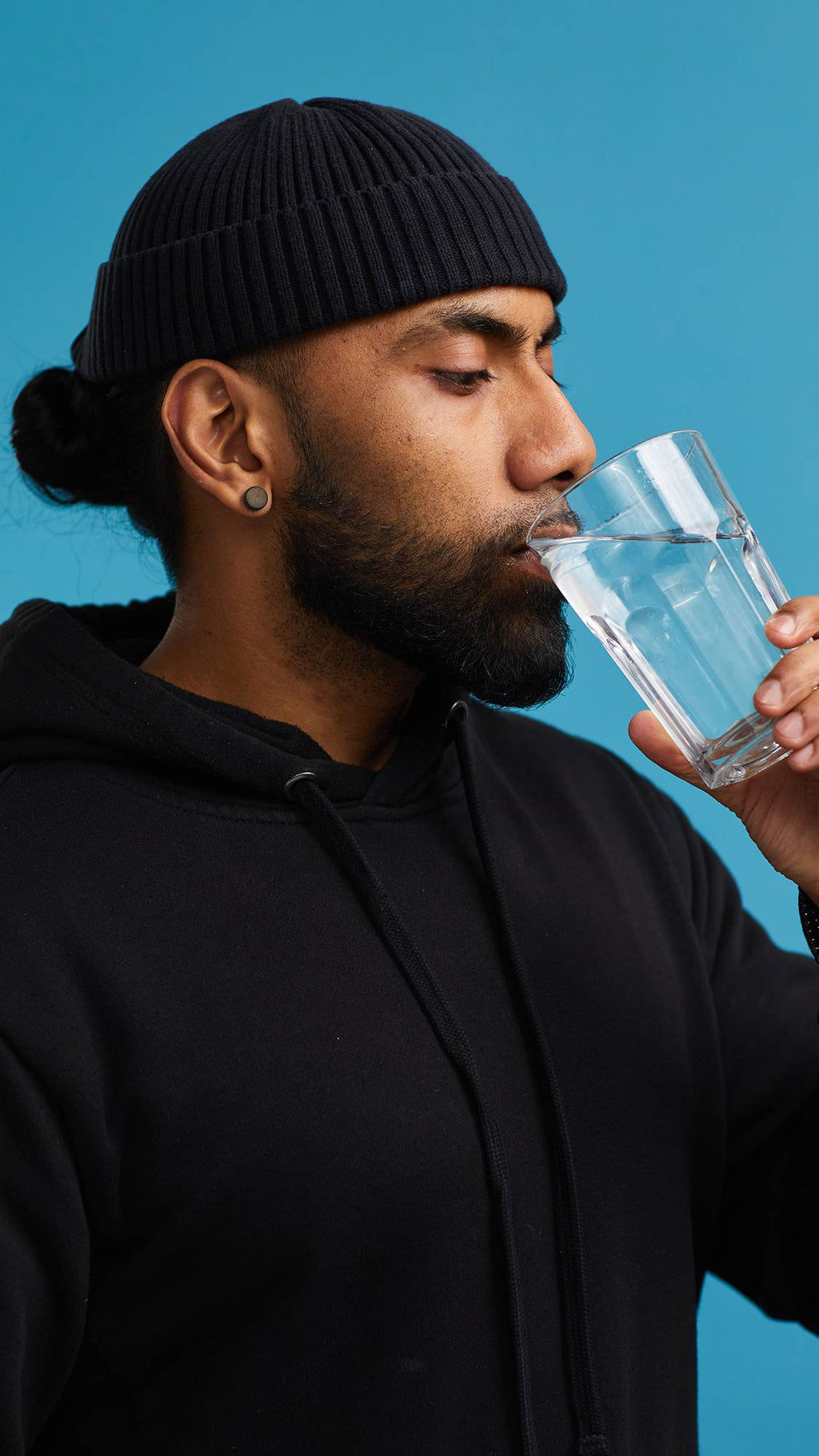 The image shows the model on a sky blue background wearing a back hoodie and hat. They have just placed one of the small, pink, The Kiss mouthwash tabs into their mouth and are taking a sip of water from a glass.