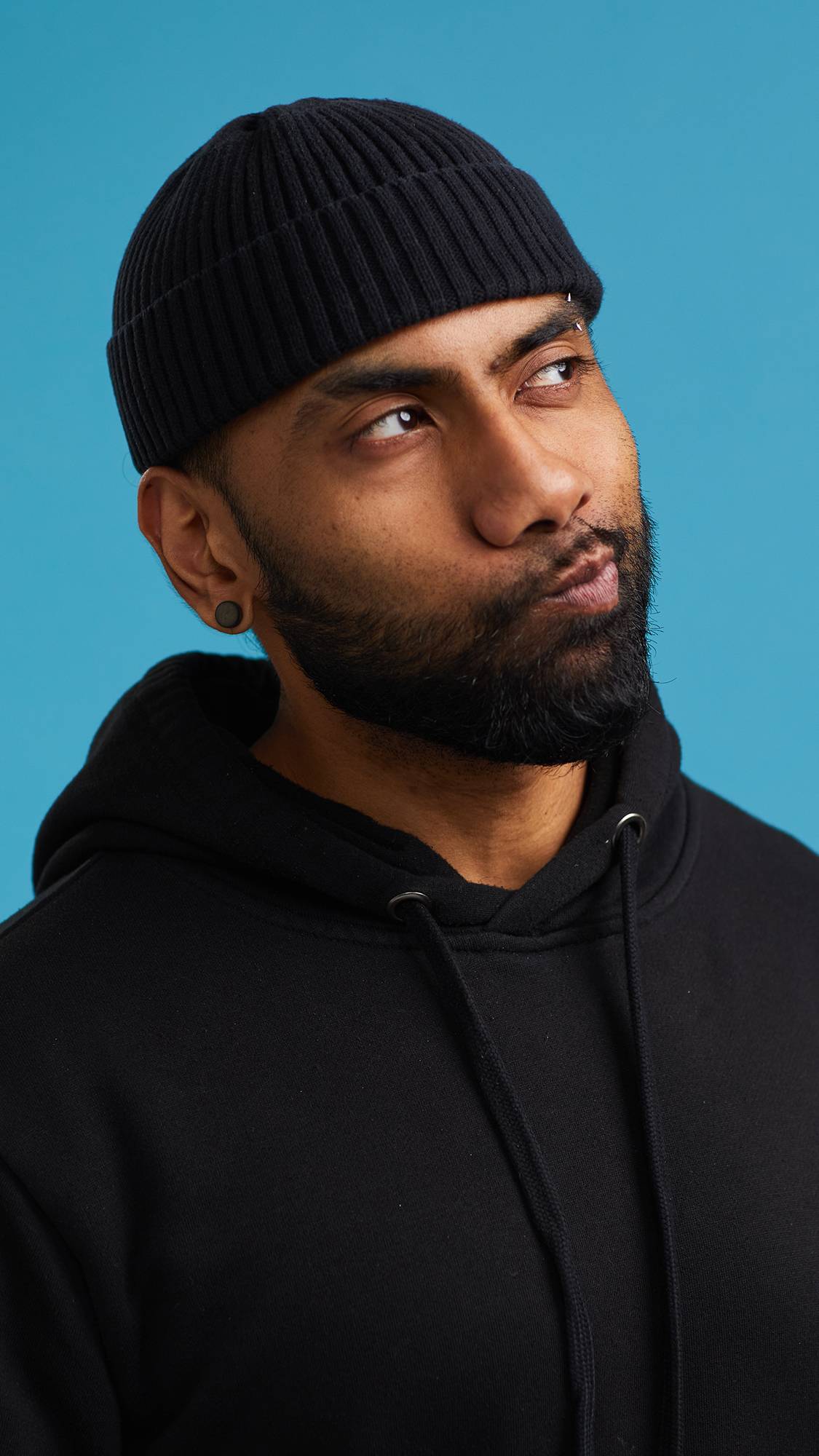 The image shows the model on a sky blue background wearing a back hoodie and hat. They have just placed one of The Kiss mouthwash tabs into their mouth and taken a mouthful of water. The image shows them now washing their mouth with water.