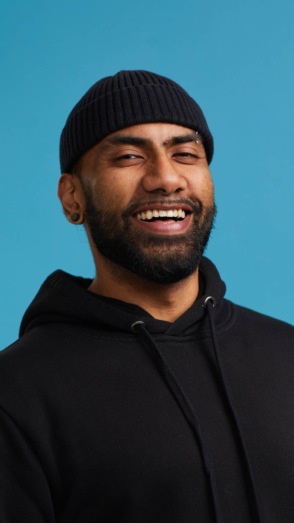 The image shows the model on a sky blue background wearing a back hoodie and hat. They have just washed their mouth with one of The Kiss mouthwash tabs and some water and are smiling at the camera with gleaming, shiny, fresh teeth.
