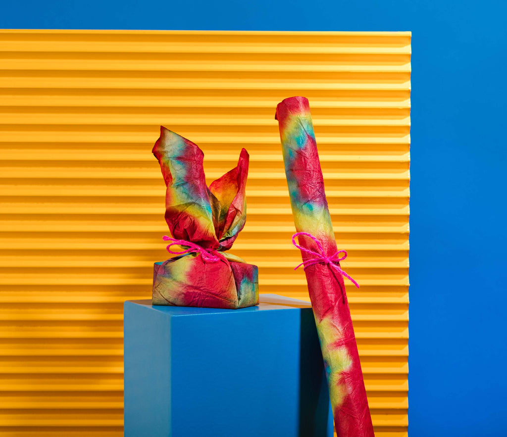 The Lokta paper is wrapped around a product, and rolled up and tied with string, on a blue plinth before an orange background.