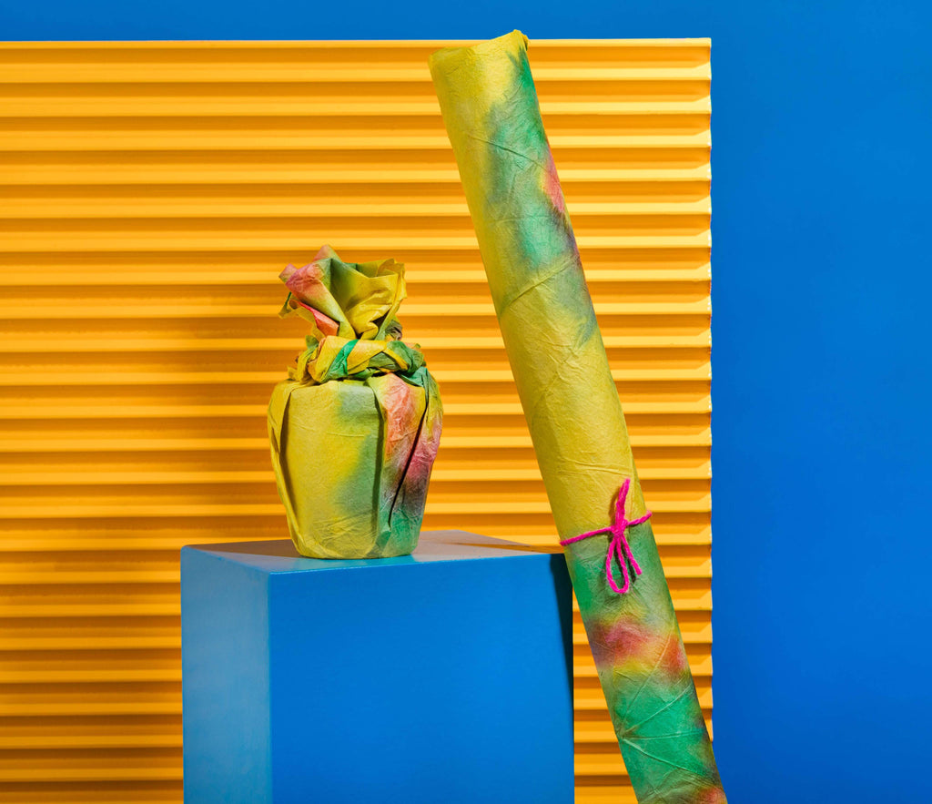 The Lokta paper is wrapped around a product, and rolled up and tied with string, on a blue plinth before an orange background.