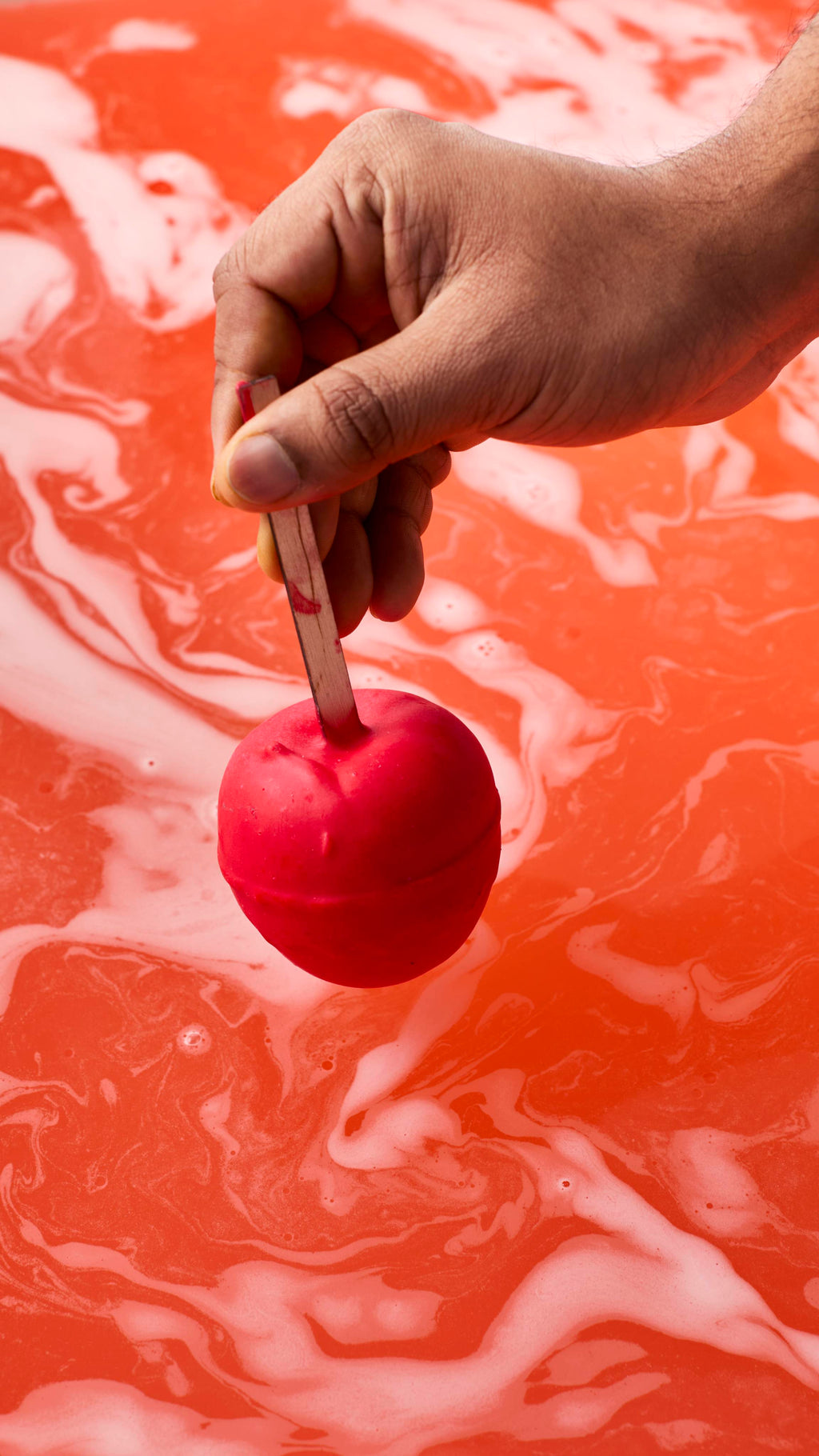 A hand holds a matte red, apple-shaped bath bomb by its wooden stick just above the water's surface. Below, the bath has turned a vibrant, opaque orange-red, covered in marbling swirls of thick white foam.
