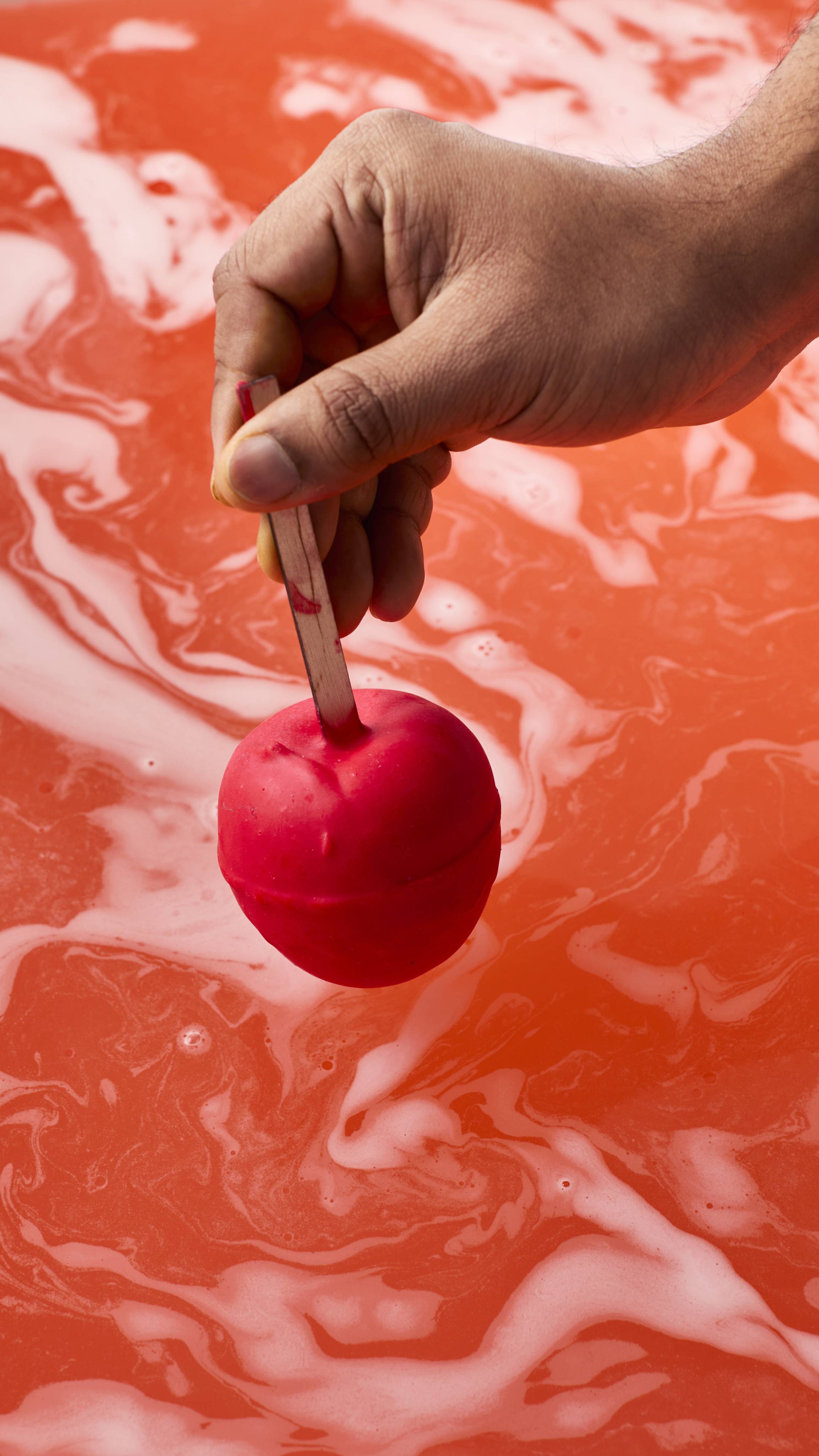 A hand holds a matte red, apple-shaped bath bomb by its wooden stick just above the water's surface. Below, the bath has turned a vibrant, opaque orange-red, covered in marbling swirls of thick white foam.