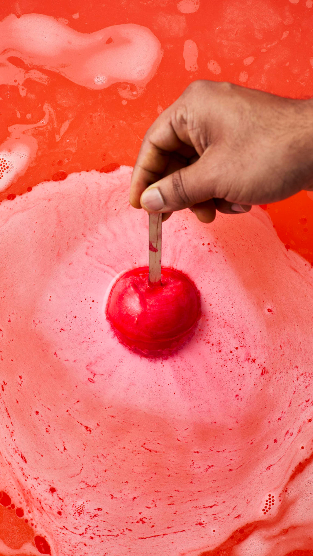 A hand holds a matte red, apple-shaped bath bomb by its wooden stick, just about to dip below the water's surface. Below, the bath has turned a vibrant, opaque orange-red, covered in marbling swirls of thick white foam.