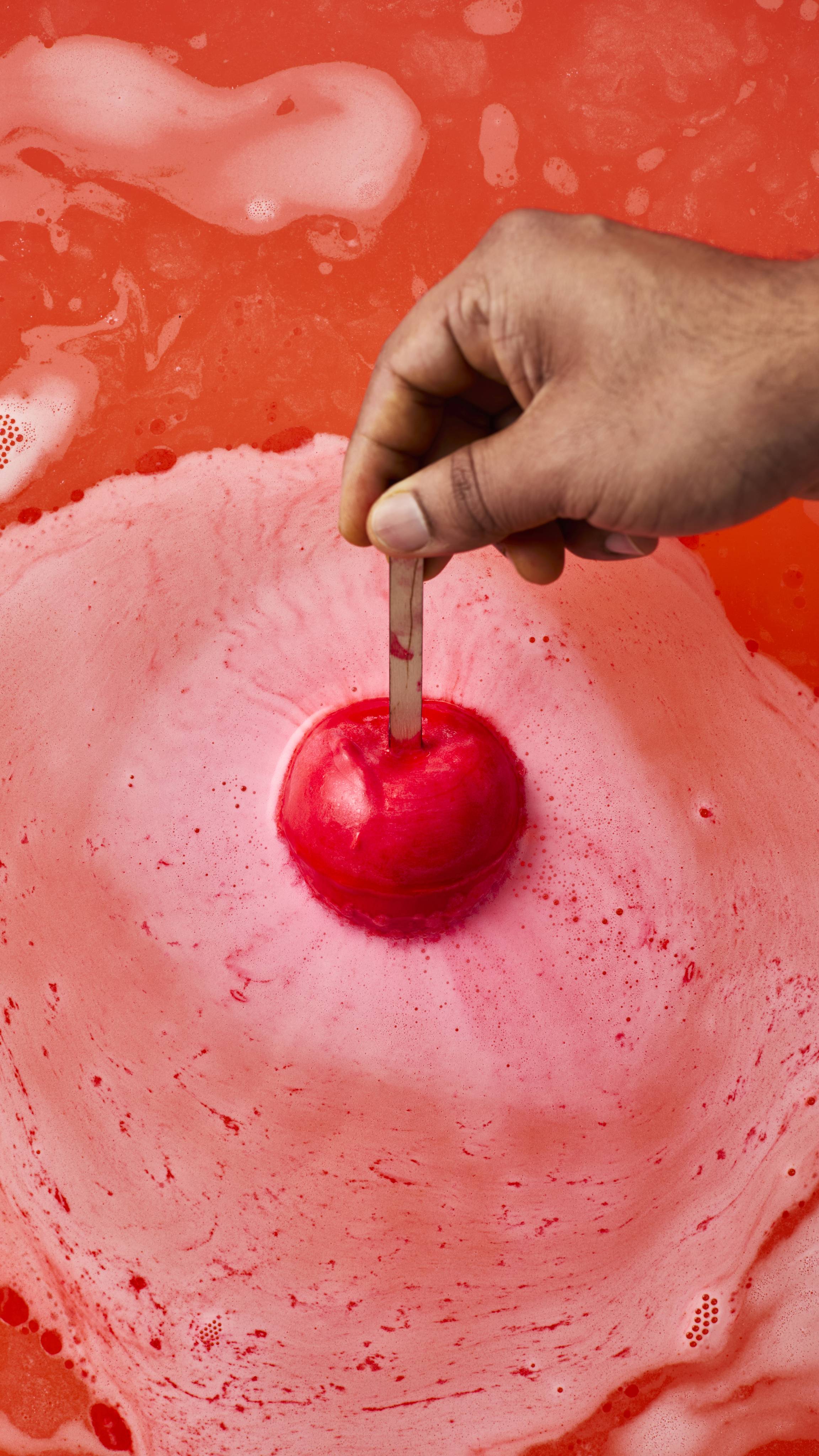 A hand holds a matte red, apple-shaped bath bomb by its wooden stick, just about to dip below the water's surface. Below, the bath has turned a vibrant, opaque orange-red, covered in marbling swirls of thick white foam.