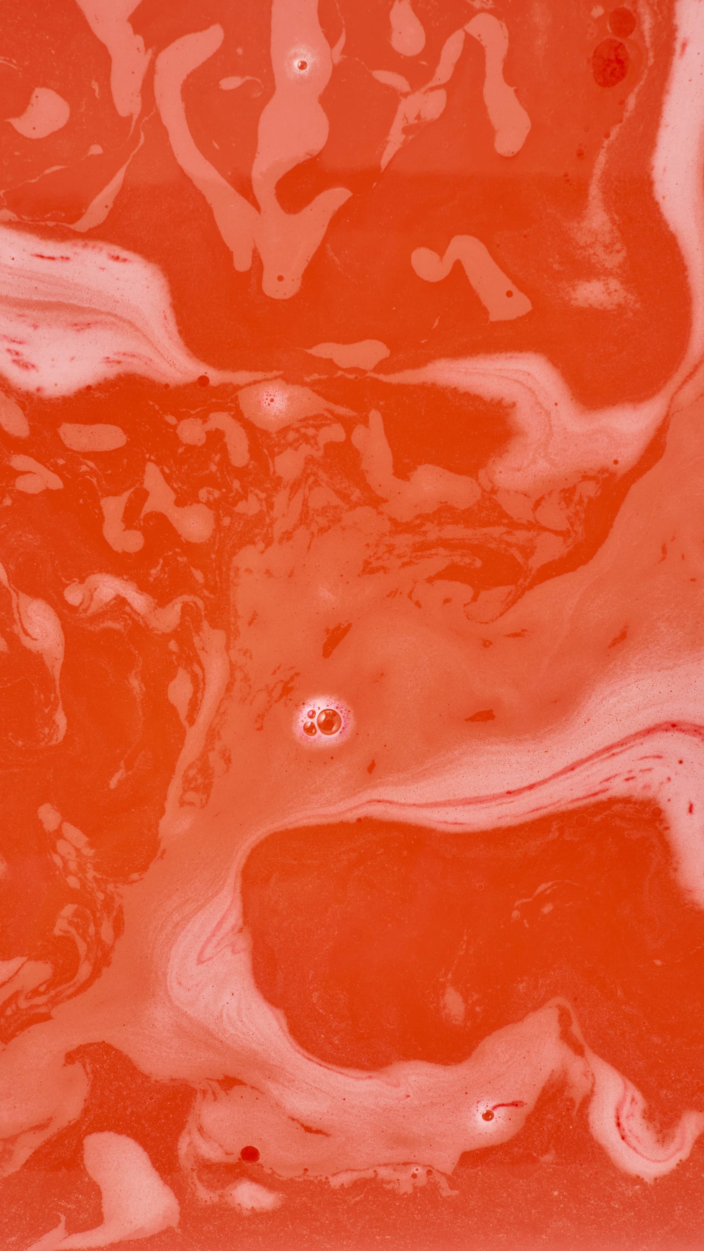 An abstract aerial shot of bath bomb foam swirling in water, creating marbled, wavy patterns of vibrant orange-red and pale pink-white froth. A few small clusters of bubbles are scattered across the colourful surface.