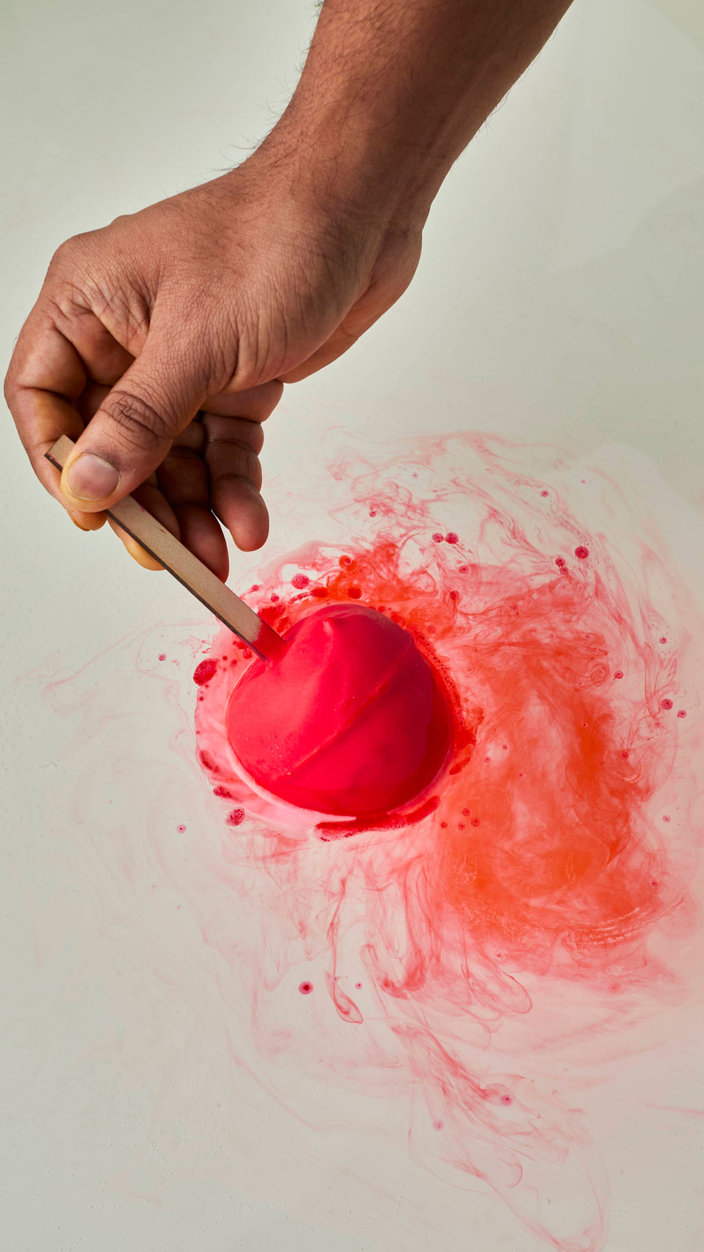 A person's hand holds a matte red, apple-shaped bath bomb by its wooden stick as it dissolves in water. It releases vibrant, swirling trails of red and orange colour across the water's surface.
