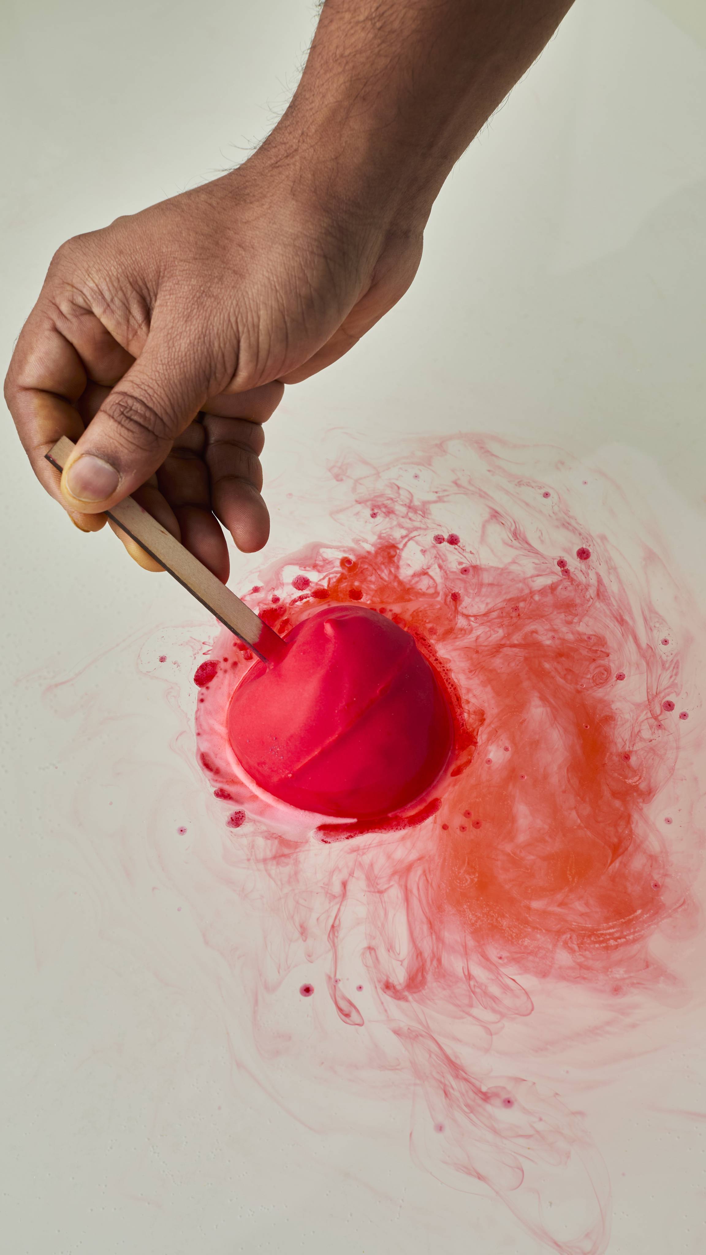 A person's hand holds a matte red, apple-shaped bath bomb by its wooden stick as it dissolves in water. It releases vibrant, swirling trails of red and orange colour across the water's surface.