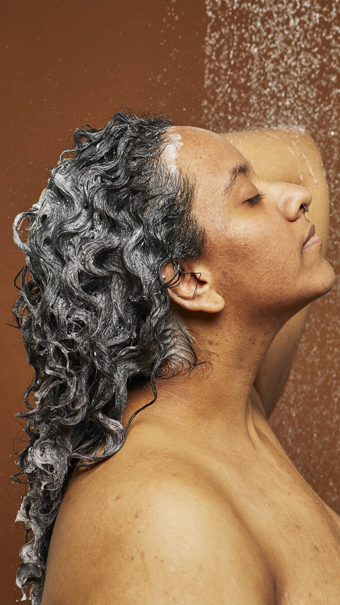 Model is standing under shower water lathering up the shampoo as it falls down their back on a deep earth-tone background.