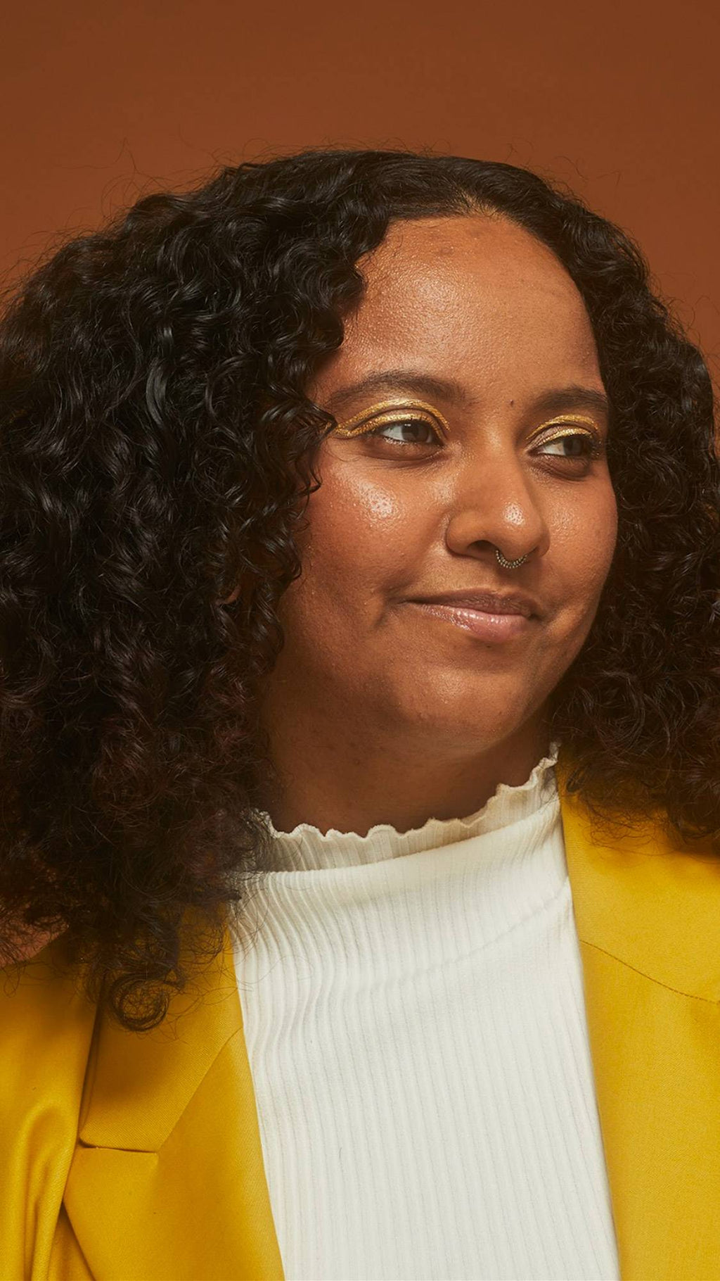 Model wears a bright yellow jacket and bold gold eyeliner with thick, defined curly dark hair on an earth-toned background.
