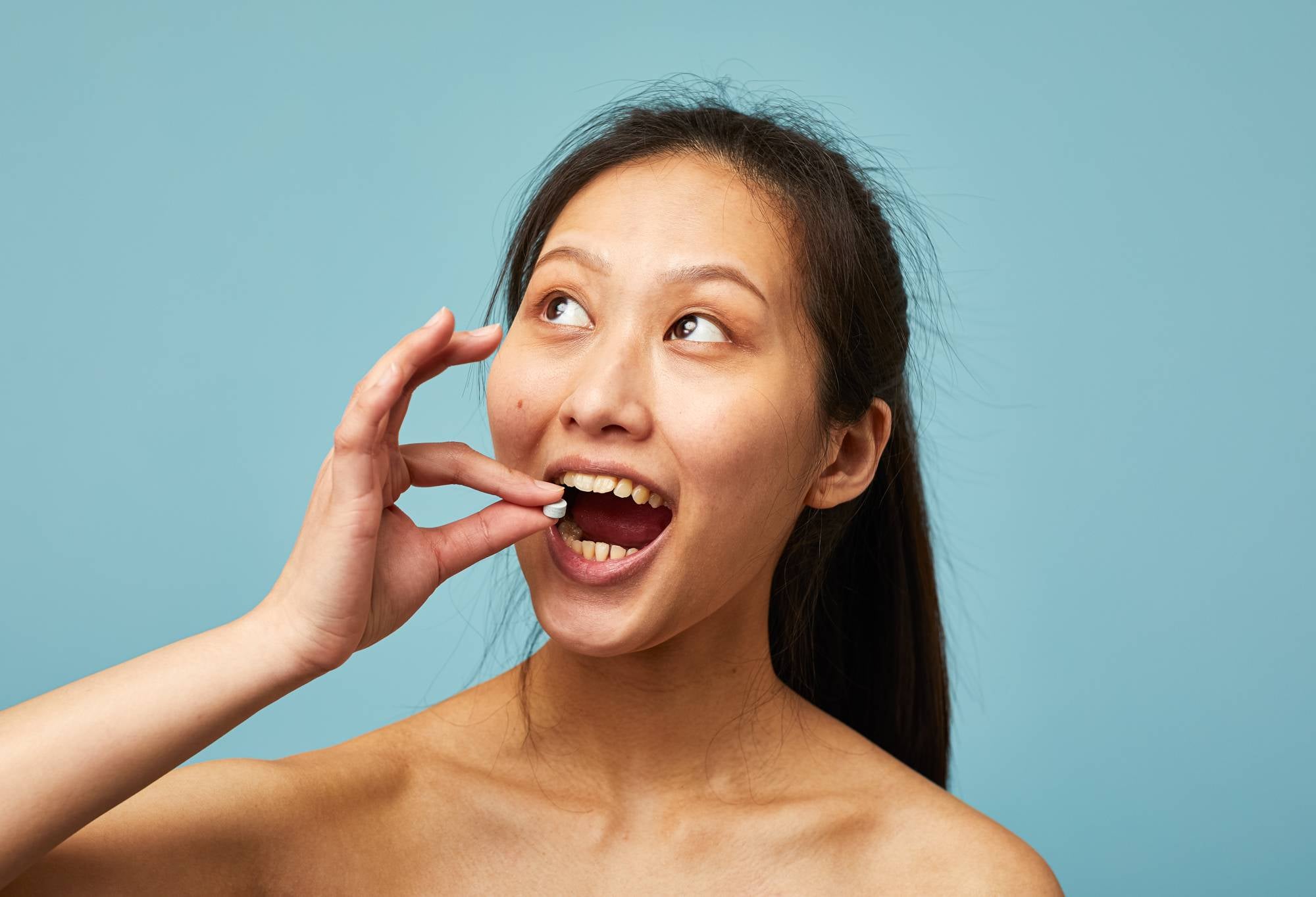 A person prepares to put a small blue toothpaste tab into their open mouth with their hand.
