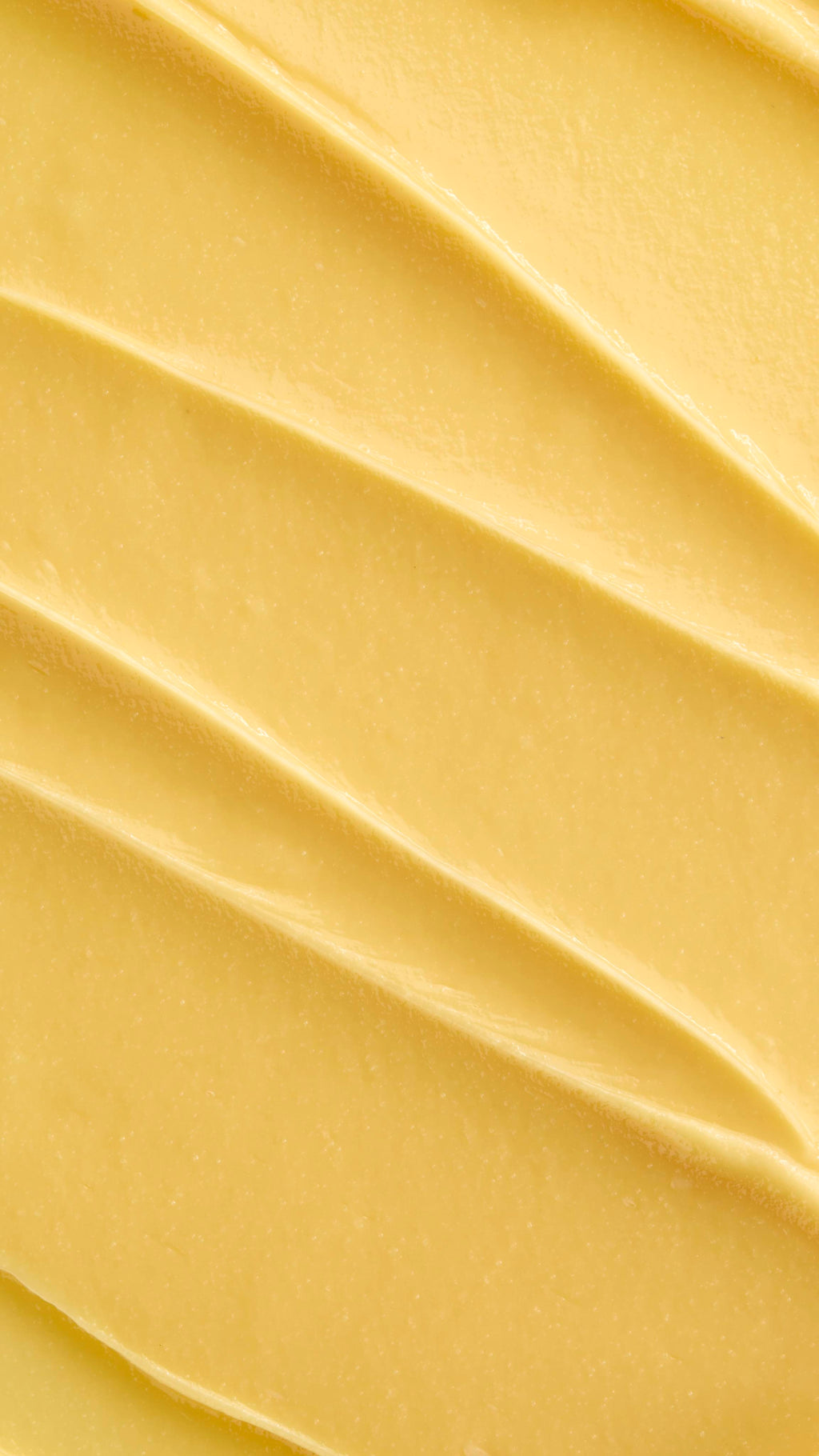 A super close-up image showing diagonal, overlapping smears of the thick, creamy, pale yellow Lush "Turmeric And Argan" facial cleanser. The smooth product has a matte finish and subtle ridged textures.