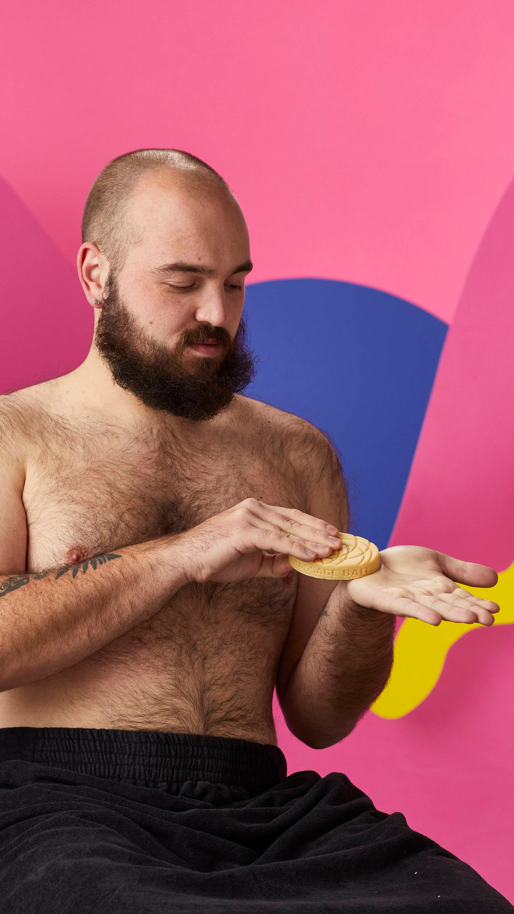 A shirtless, bearded person wrapped in a black towel holds an oval Lush massage bar, warming it between their hands. Set against a vibrant background of pink, blue, and yellow abstract shapes.
