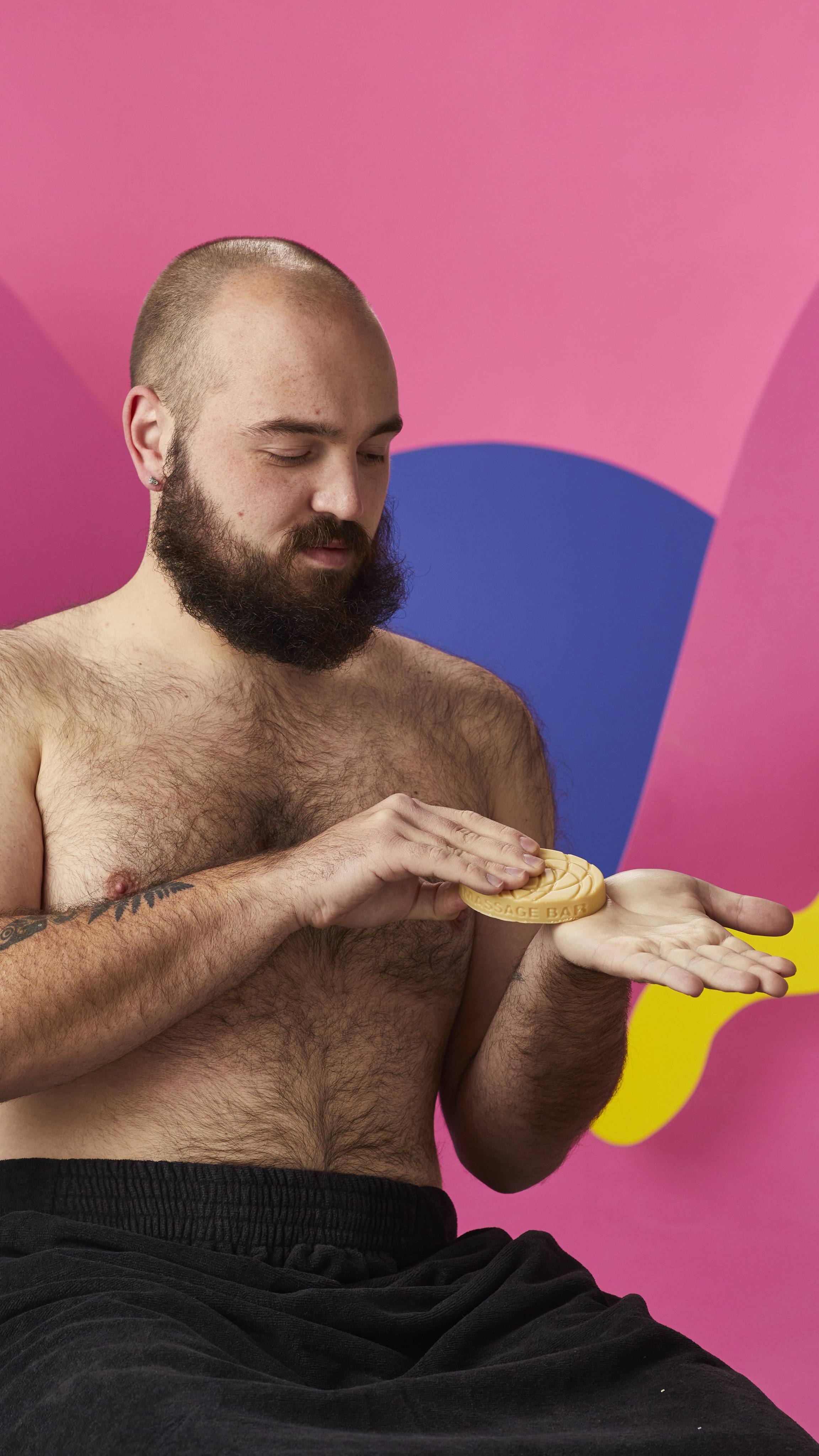 A shirtless, bearded person wrapped in a black towel holds an oval Lush massage bar, warming it between their hands. Set against a vibrant background of pink, blue, and yellow abstract shapes.