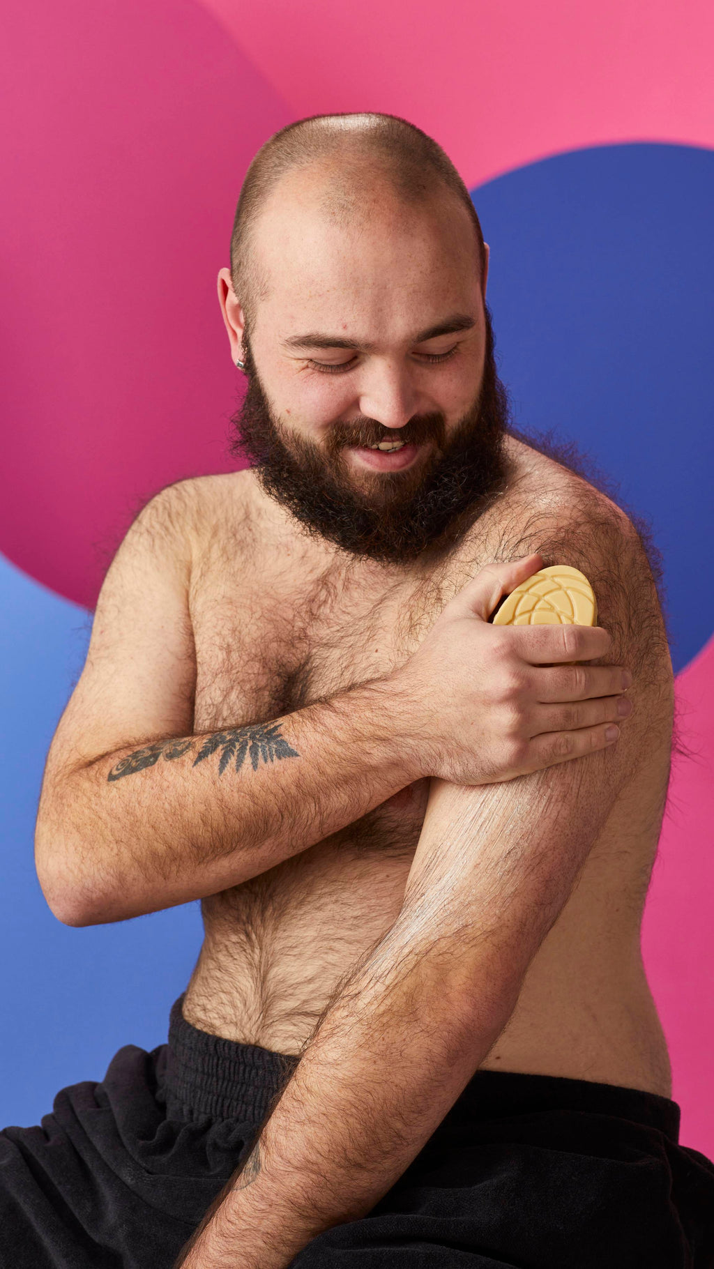 A smiling, shirtless bearded person sweeps a smooth, patterned Lush massage bar down their upper arm, leaving a moisturizing sheen. They are wrapped in a black towel against a vibrant, abstract background of pink and blue curves.