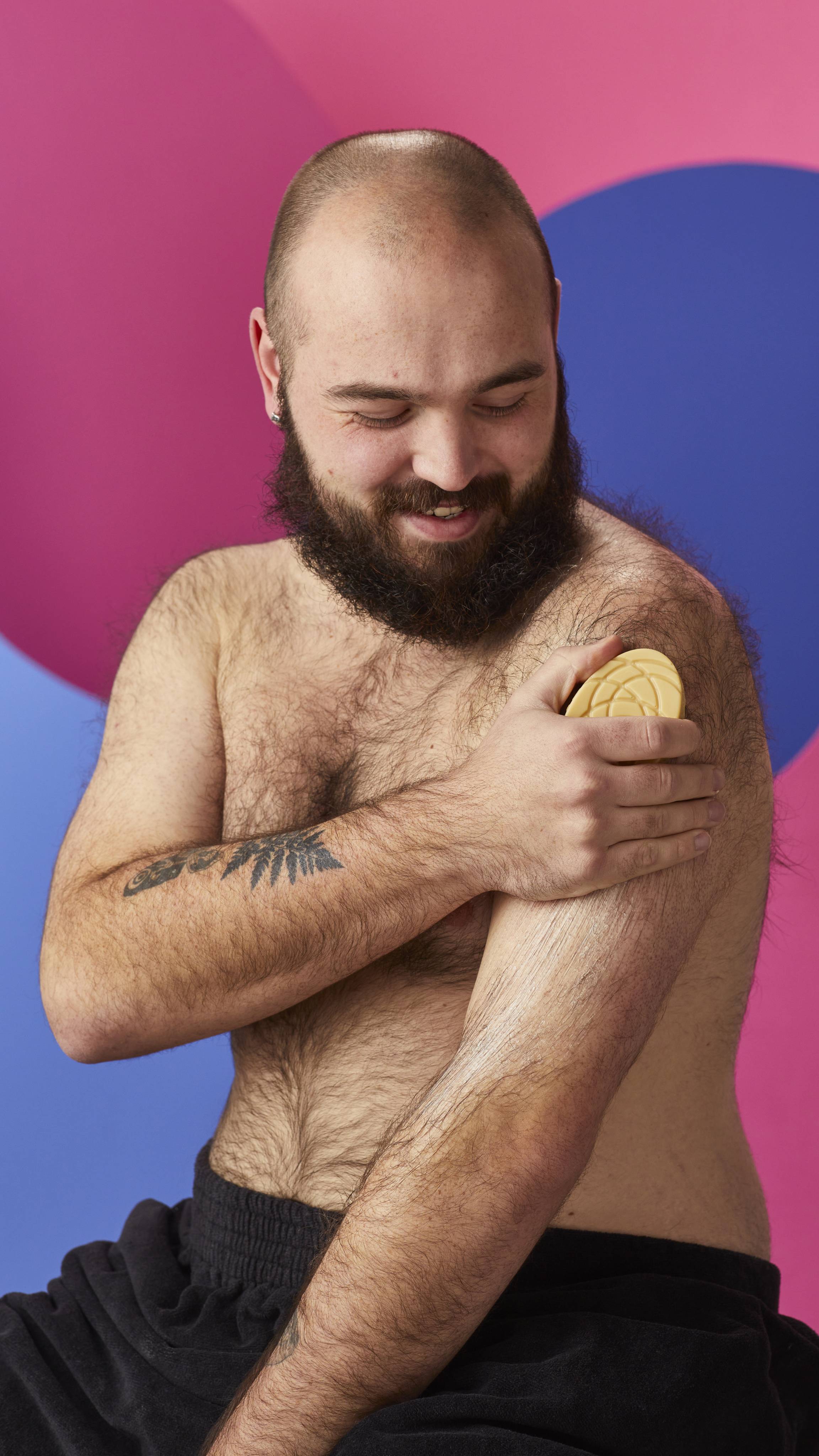 A smiling, shirtless bearded person sweeps a smooth, patterned Lush massage bar down their upper arm, leaving a moisturizing sheen. They are wrapped in a black towel against a vibrant, abstract background of pink and blue curves.