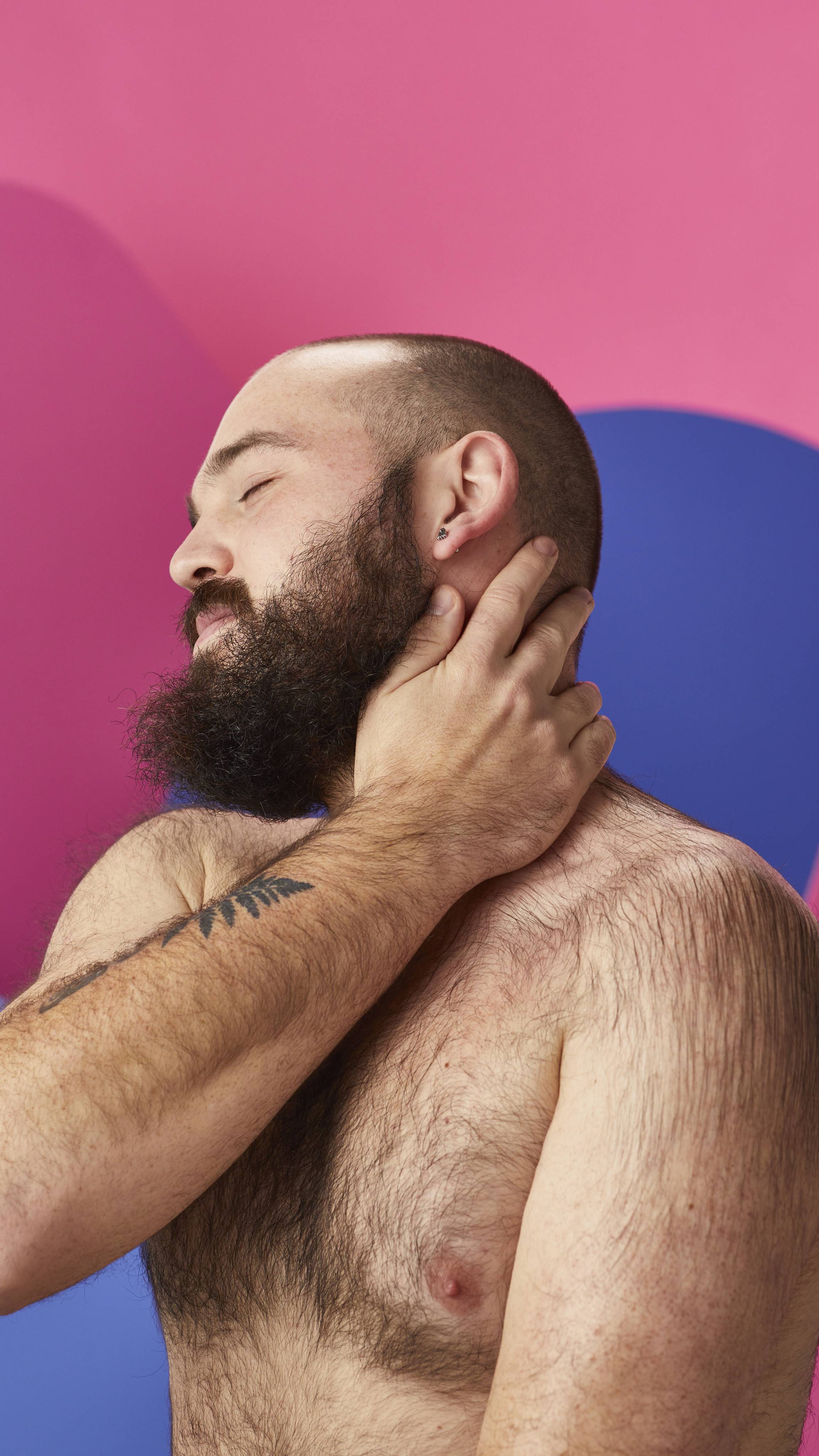 A shirtless, bearded person relaxes with eyes closed, gently massaging their neck with one hand to absorb the moisturizing oils from a Lush massage bar. Set against a vibrant abstract background of bold pink and blue shapes.
