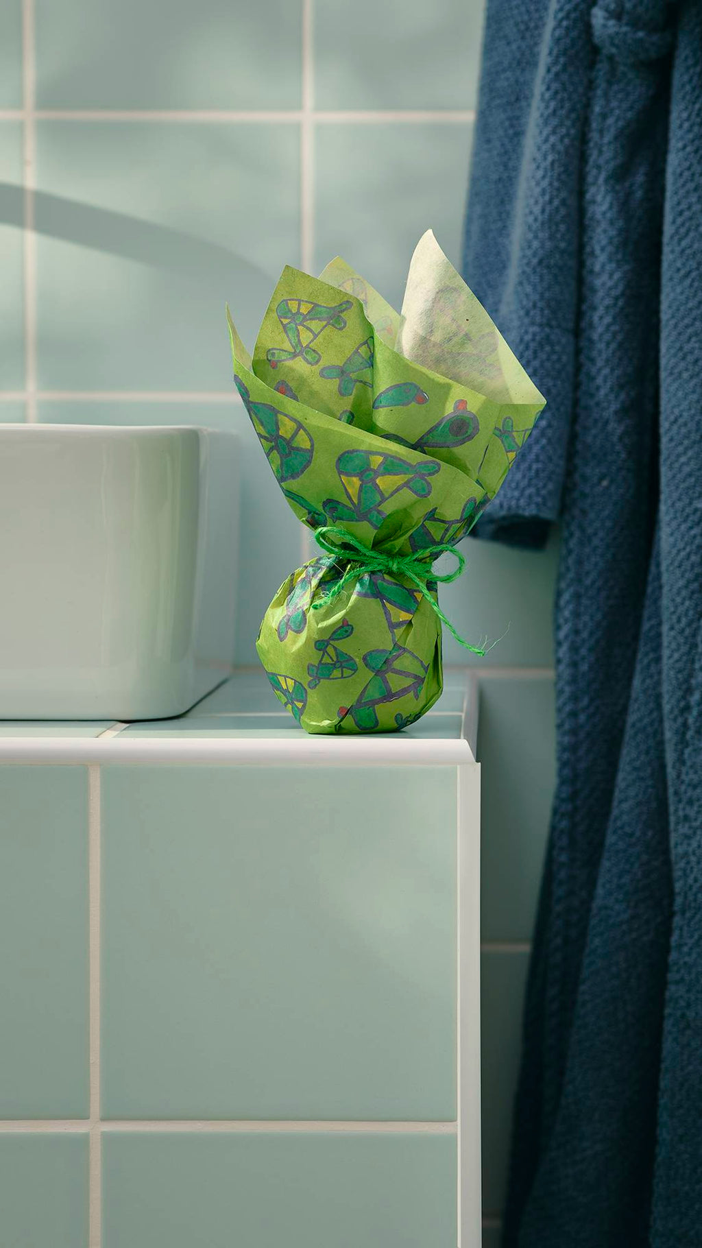 A small, spherical gift, wrapped in green "Turtley Awesome" Lokta paper with a hand-drawn turtle pattern, sits on a tiled bathroom ledge. The paper is gathered at the top and tied with twine. A dressing gown hangs in the background.