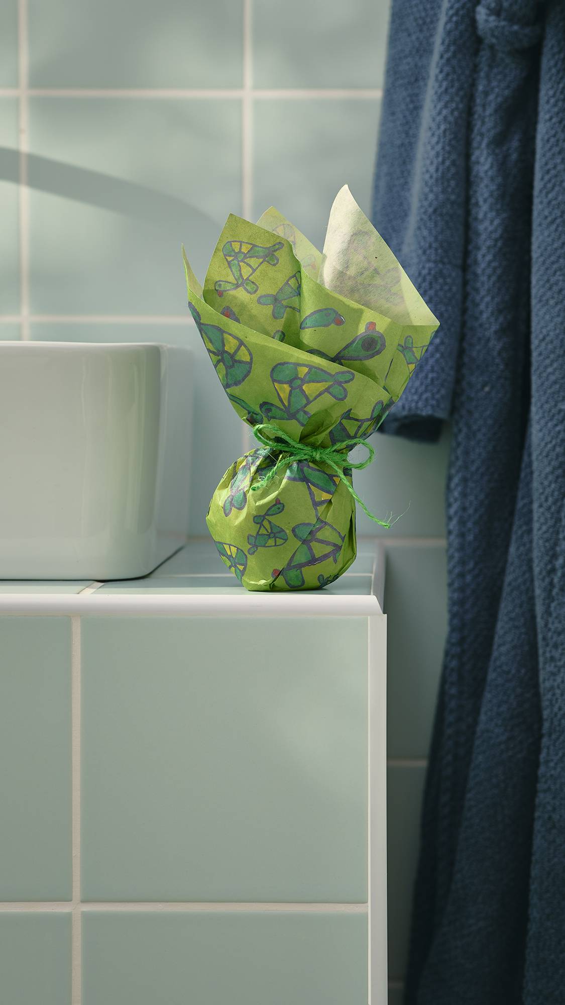 A small, spherical gift, wrapped in green "Turtley Awesome" Lokta paper with a hand-drawn turtle pattern, sits on a tiled bathroom ledge. The paper is gathered at the top and tied with twine. A dressing gown hangs in the background.