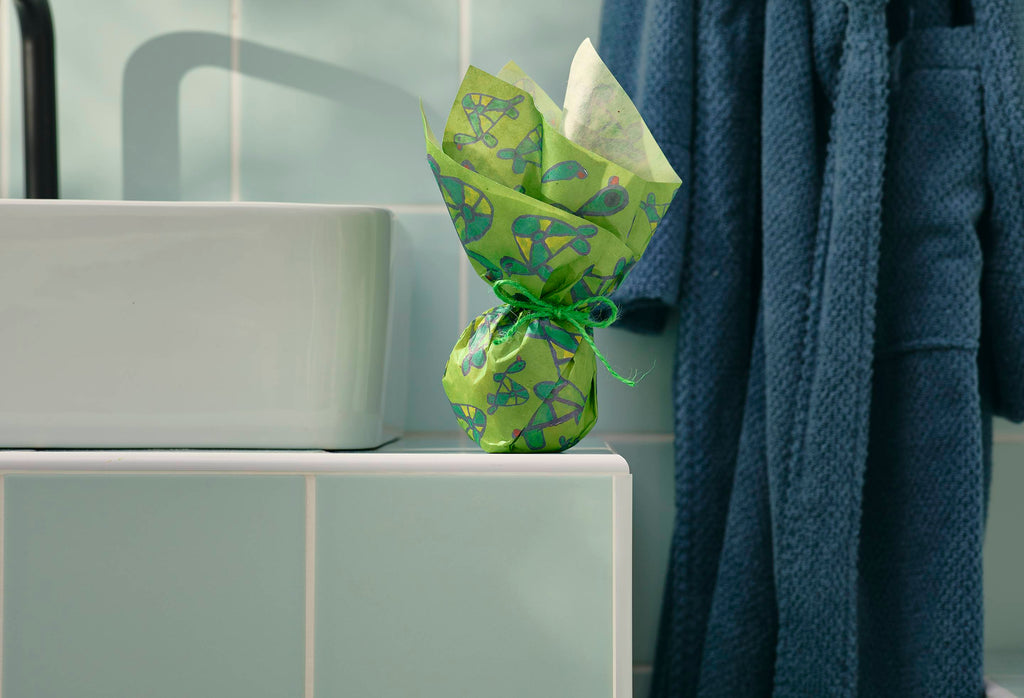 A small, spherical gift, wrapped in green "Turtley Awesome" Lokta paper with a hand-drawn turtle pattern, sits on a tiled bathroom ledge. The paper is gathered at the top and tied with twine. A dressing gown hangs in the background.
