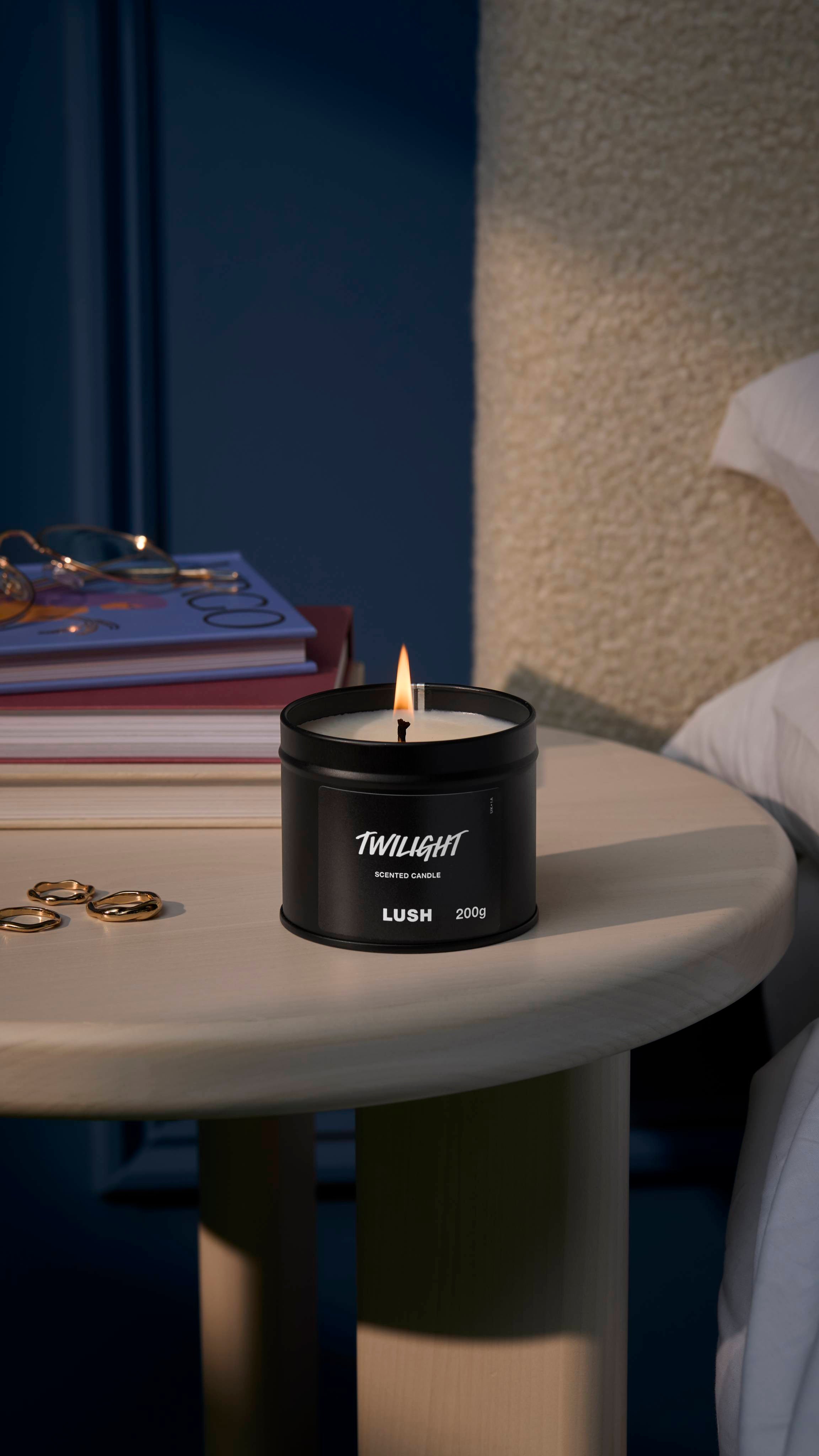 The image shows a deep, midnight-blue walled background with a cream bed headboard just visible. The black tin Twilight candle sits lit on a white, wooden side table by a small pile of books, glasses, and jewellery.