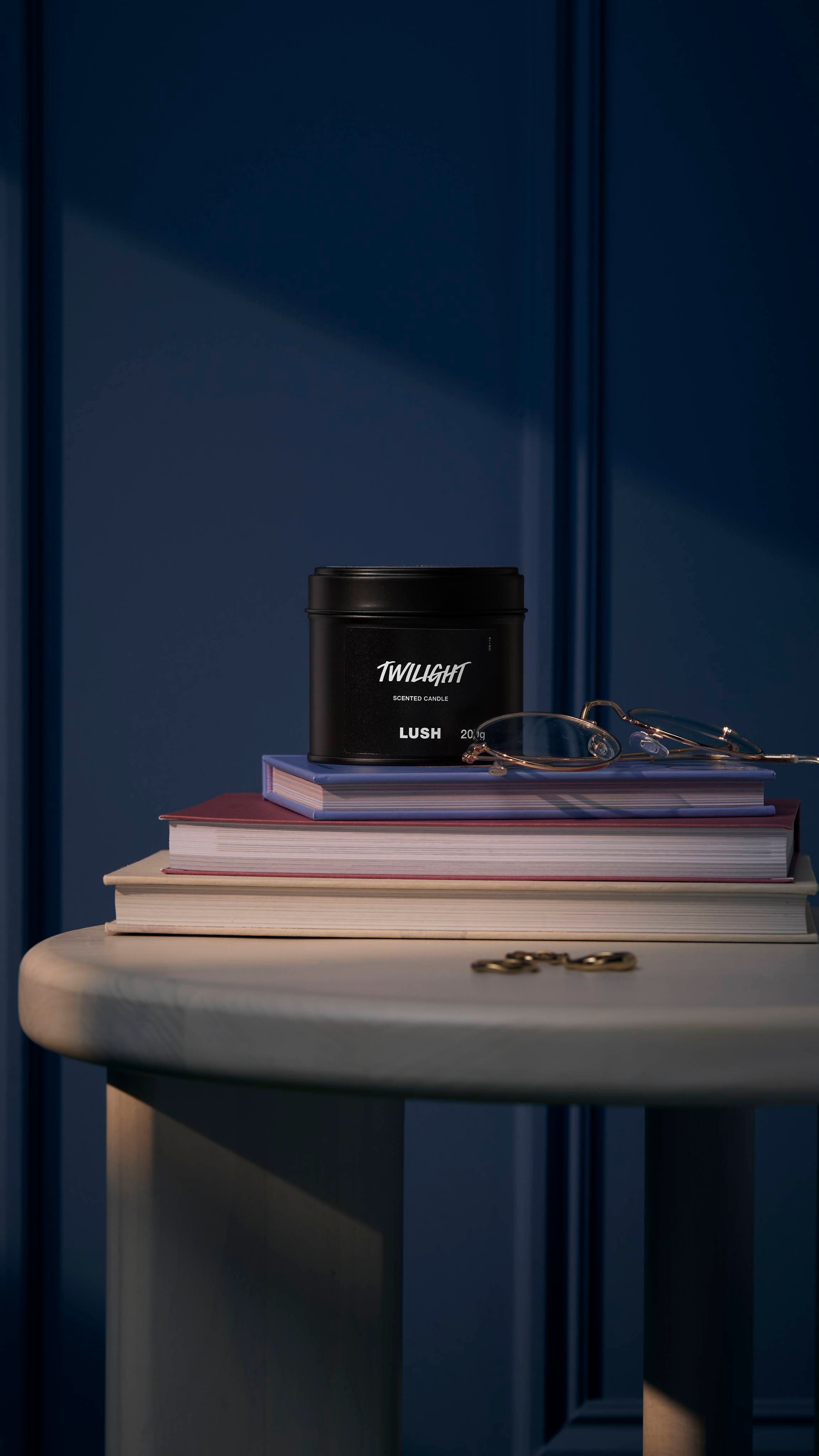 The image shows a deep, midnight-blue walled background. The black tin Twilight candle sits on a white, wooden side table on top of a small pile of books, glasses, and jewellery.