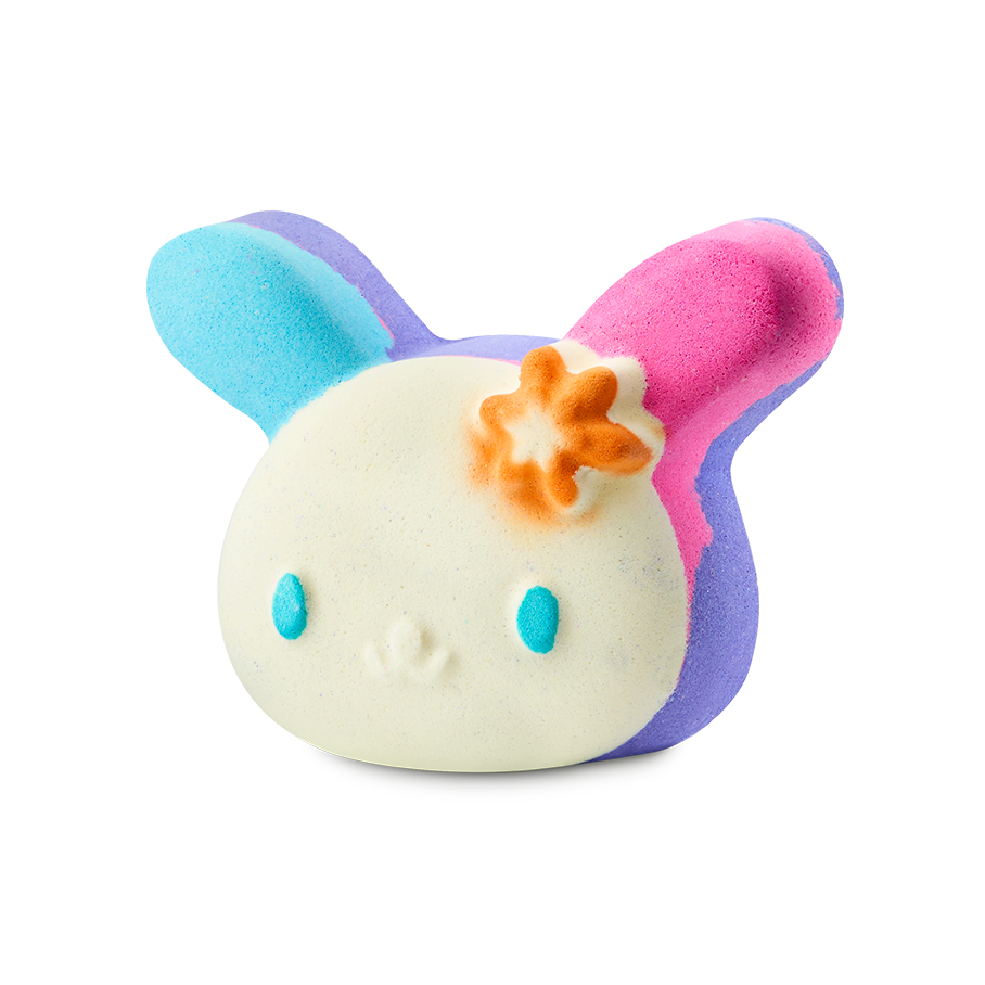U.SA.HA.NA. A colourful bath bomb shaped like a bunny character's head with a cream face, blue eyes, and an orange flower. One ear is bright blue and the other is pink, with purple accents throughout.