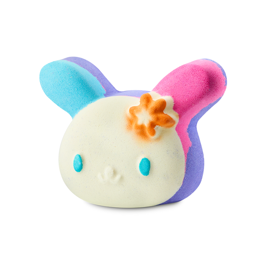 U.SA.HA.NA. A colourful bath bomb shaped like a bunny character's head with a cream face, blue eyes, and an orange flower. One ear is bright blue and the other is pink, with purple accents throughout.