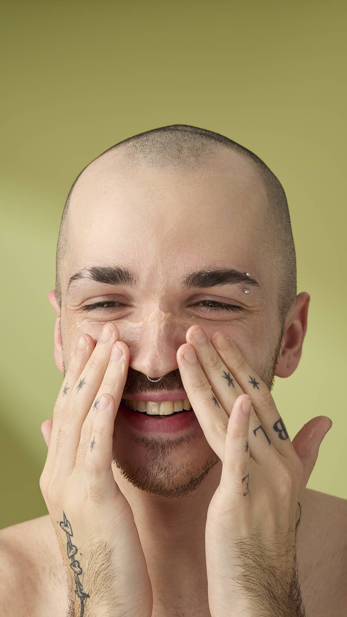 A close-up of a person with a shaved head and facial hair smiling as they massage a creamy 'Ultraplant' facial cleanser into their nose and cheeks, set against a soft olive-green background.
