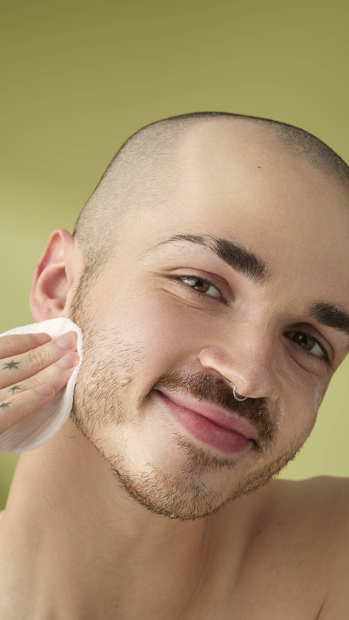 A close-up portrait of a person with a shaved head and facial hair, smiling as they wipe the 'Utlraplant' facial cleanser off their cheek with a white cotton pad, set against a soft olive-green background.