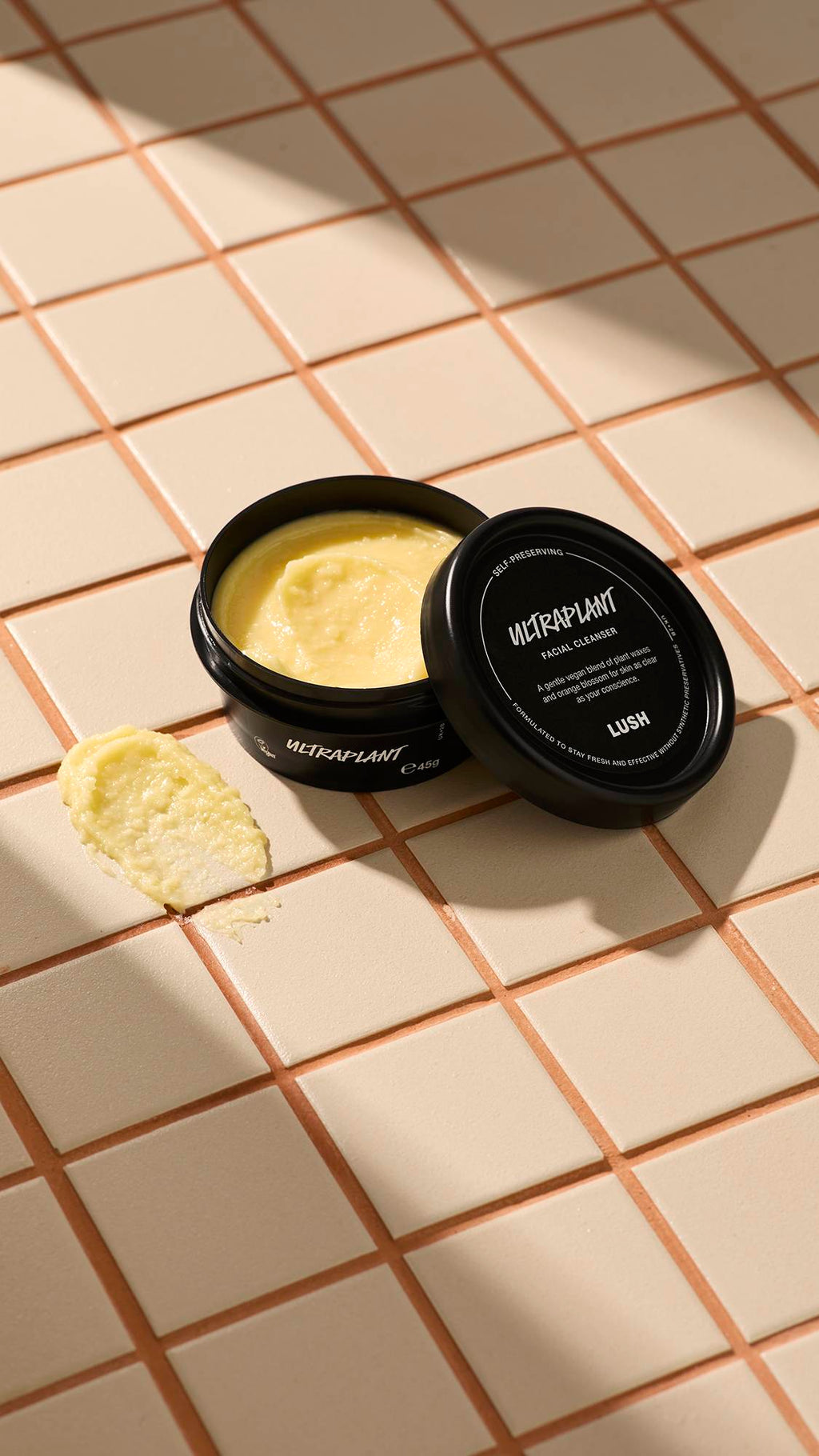 An open black tub of Lush 'Ultraplant' facial cleanser sits on a beige tiled surface. Beside it is a small smear of the thick, pale yellow product. The lid rests against the pot, showing the product name and Lush logo in white text.