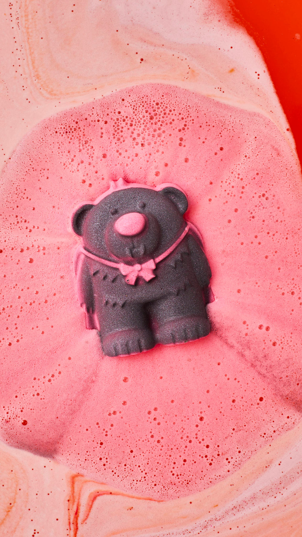 A dark grey vampire bear bath bomb with a pink bow fizzes and dissolves in bathwater, creating a large pool of frothy pink foam surrounded by orange and peach-coloured swirls.